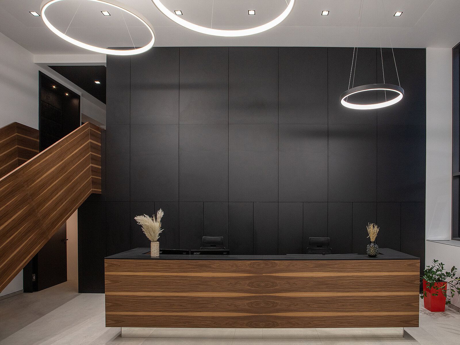 Eine moderne Empfangstheke aus Holz mit einer eleganten, dunklen Wand im Hintergrund. Über der Theke hängen kreisförmige Deckenleuchten. Daneben sind grüne Pflanzen und eine Treppe sichtbar. Die Atmosphäre ist schlicht und zeitgemäß.