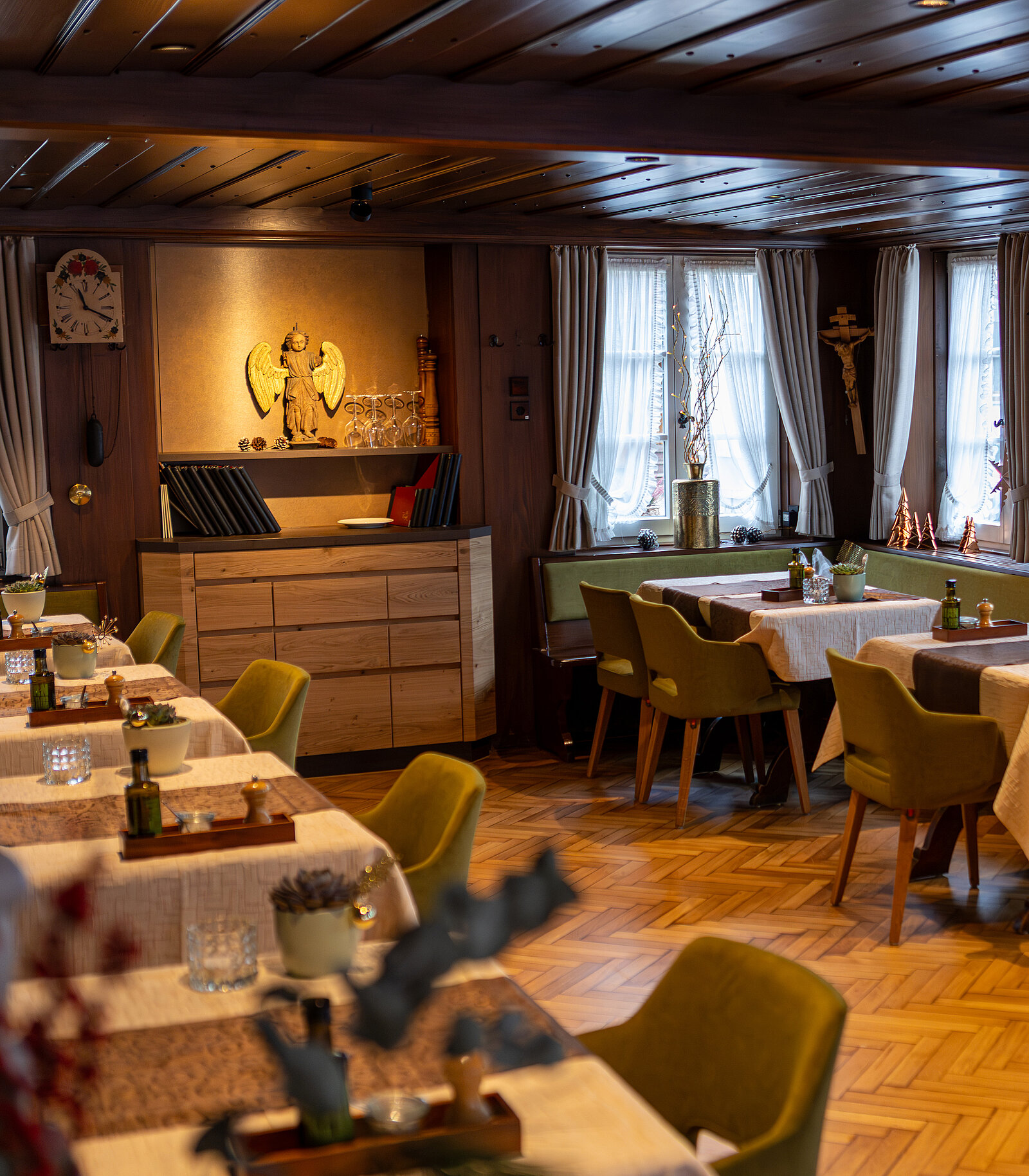 Gemütliches Restaurant mit Holzboden und -decke, dekoriert mit Engelsskulptur und Kreuzen. Tische sind mit weißen Tischdecken und Stühlen in Grün eingerichtet. Große Fenster lassen Tageslicht herein, schaffen eine warme Atmosphäre.