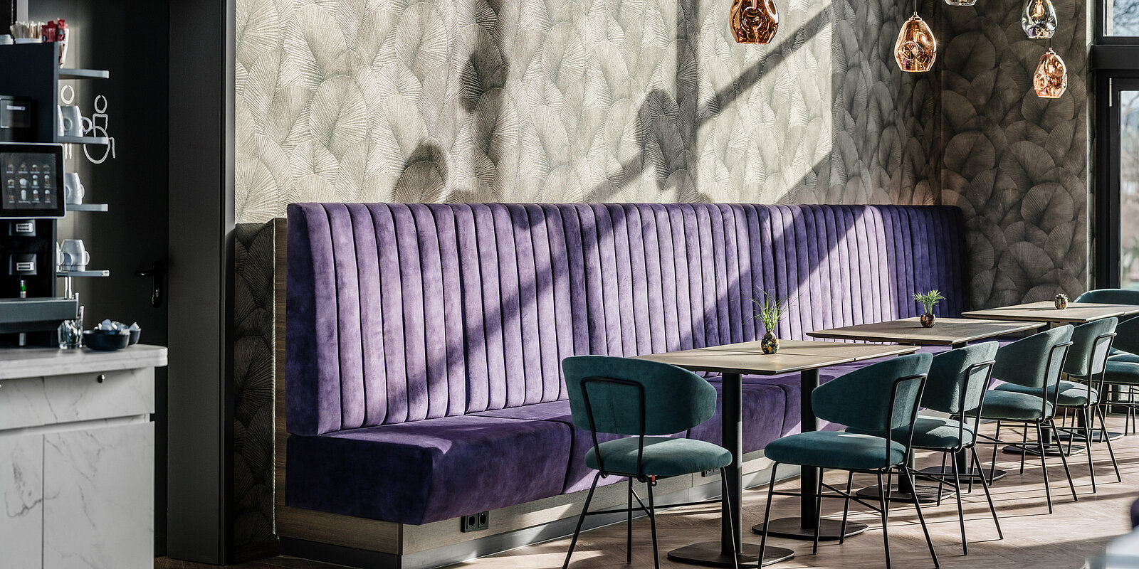 Ein stilvoll eingerichteter Cafébereich mit einer lila gepolsterten Bank, modernen Stühlen und Tischen. An der Wand hängen dekorative Lampen, und die Wand hat ein dezentes, elegantes Muster. Sonneneinstrahlung erzeugt einen gemütlichen und einladenden Eindruck.