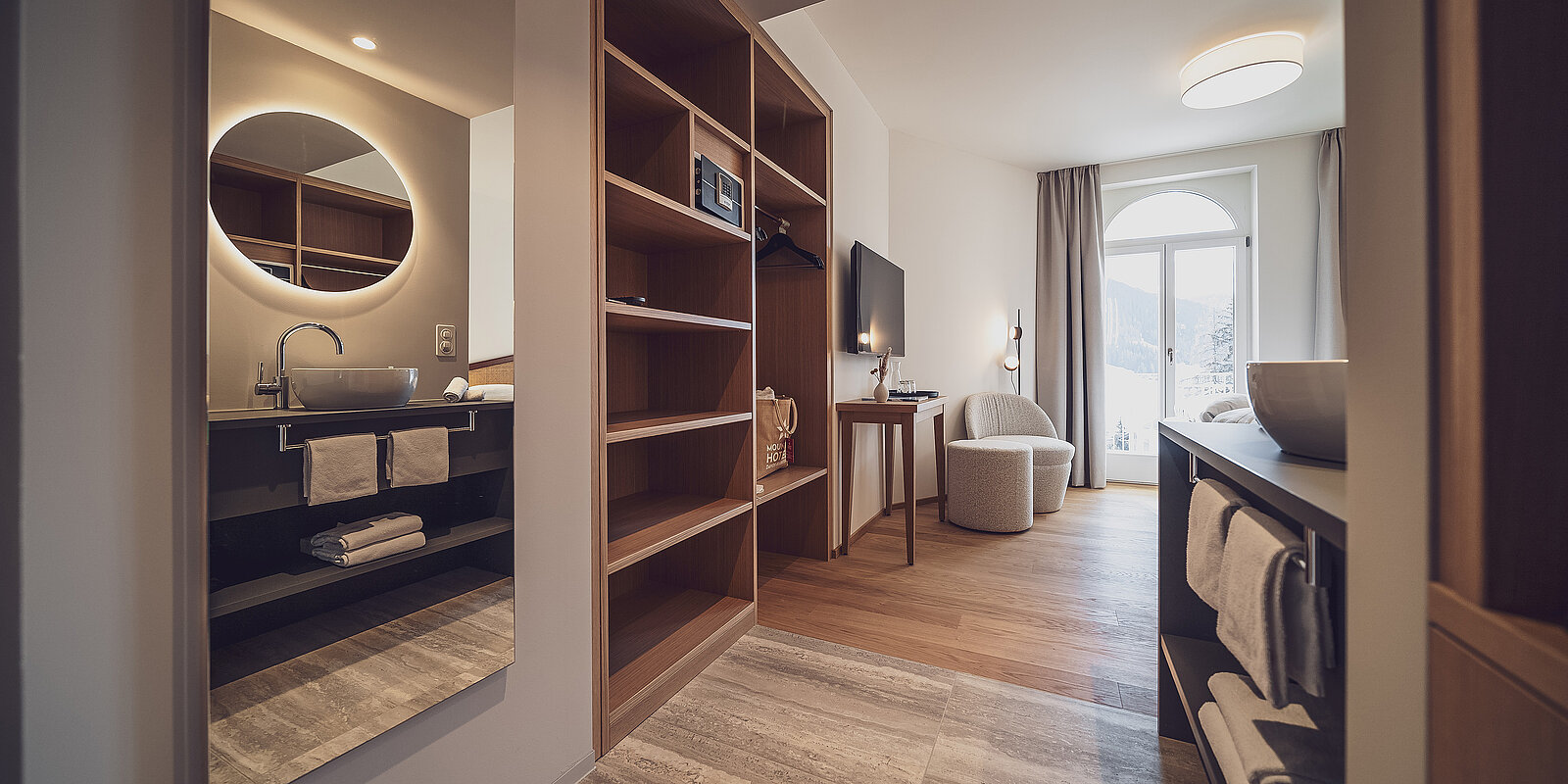 Modernes Hotelzimmer mit hellem Holzboden und stilvollen Möbeln. Links ein elegantes Badezimmer mit rundem Spiegel und Waschbecken, rechts gemütlicher Wohnbereich mit Sessel und Schreibtisch. Große Fenster lassen viel Tageslicht herein. Harmonische Atmosphäre, ideal zum Entspannen.