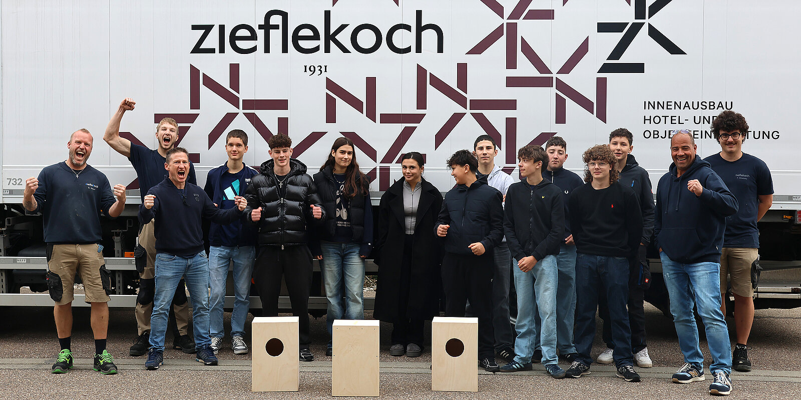 Eine Gruppe von 15 Personen steht vor einem Lastwagen, einige lächeln und jubeln. Die Männer tragen einheitliche dunkelblaue Shirts. Im Vordergrund stehen zwei kleine, quadratische Kisten. Der Lastwagen hat ein Logo und den Namen „zieflekoch 1931“ aufgedruckt.