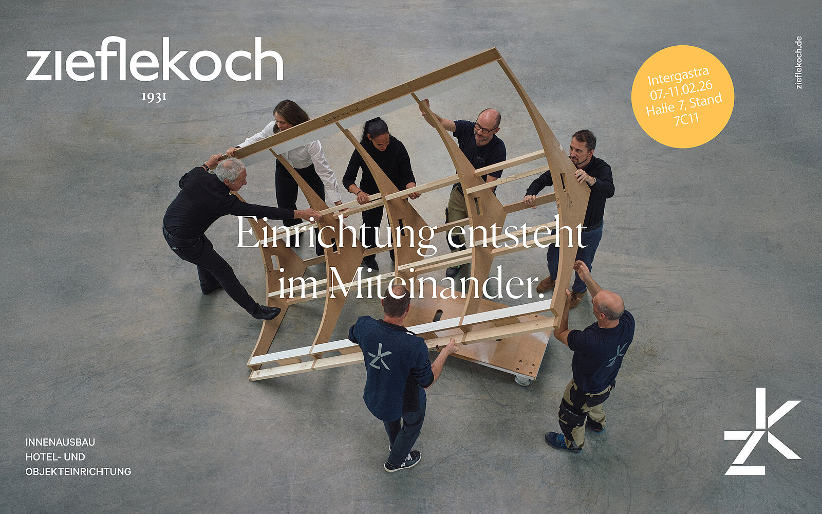 Sechs Personen bauen gemeinsam ein Holzmöbelstück auf. Der Slogan „Einrichtung entsteht im Miteinander“ ist prominent platziert. Im Hintergrund ist ein neutraler, heller Boden zu sehen.