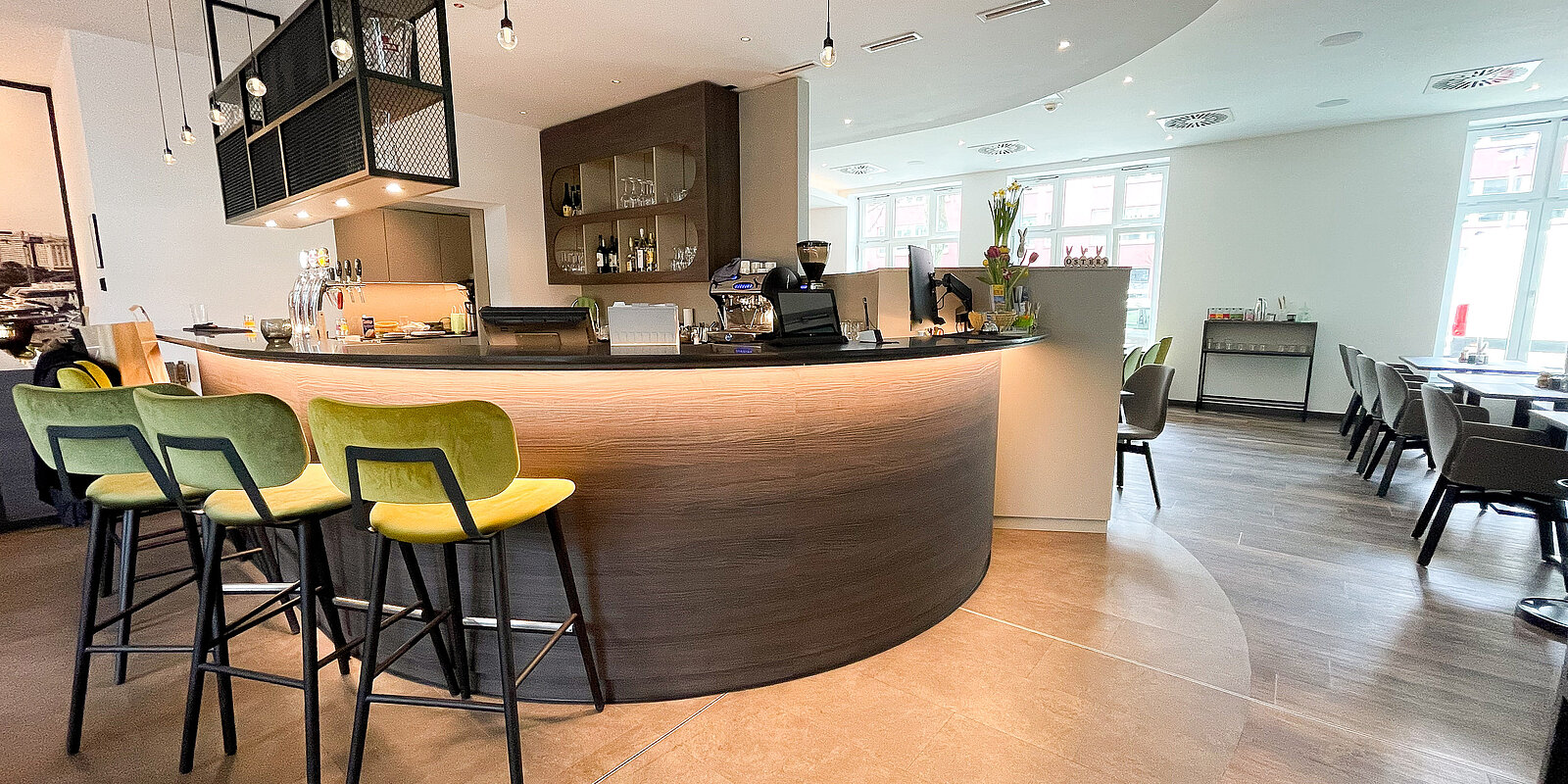 Eine moderne, geschwungene Bar mit hellen Holzoberflächen und grünen Barhockern steht zentral im Raum. Dahinter sind Regale mit Flaschen und Gläsern sichtbar. Rechts sind Tische und Stühle in einem offenen und hellen Essbereich angeordnet.