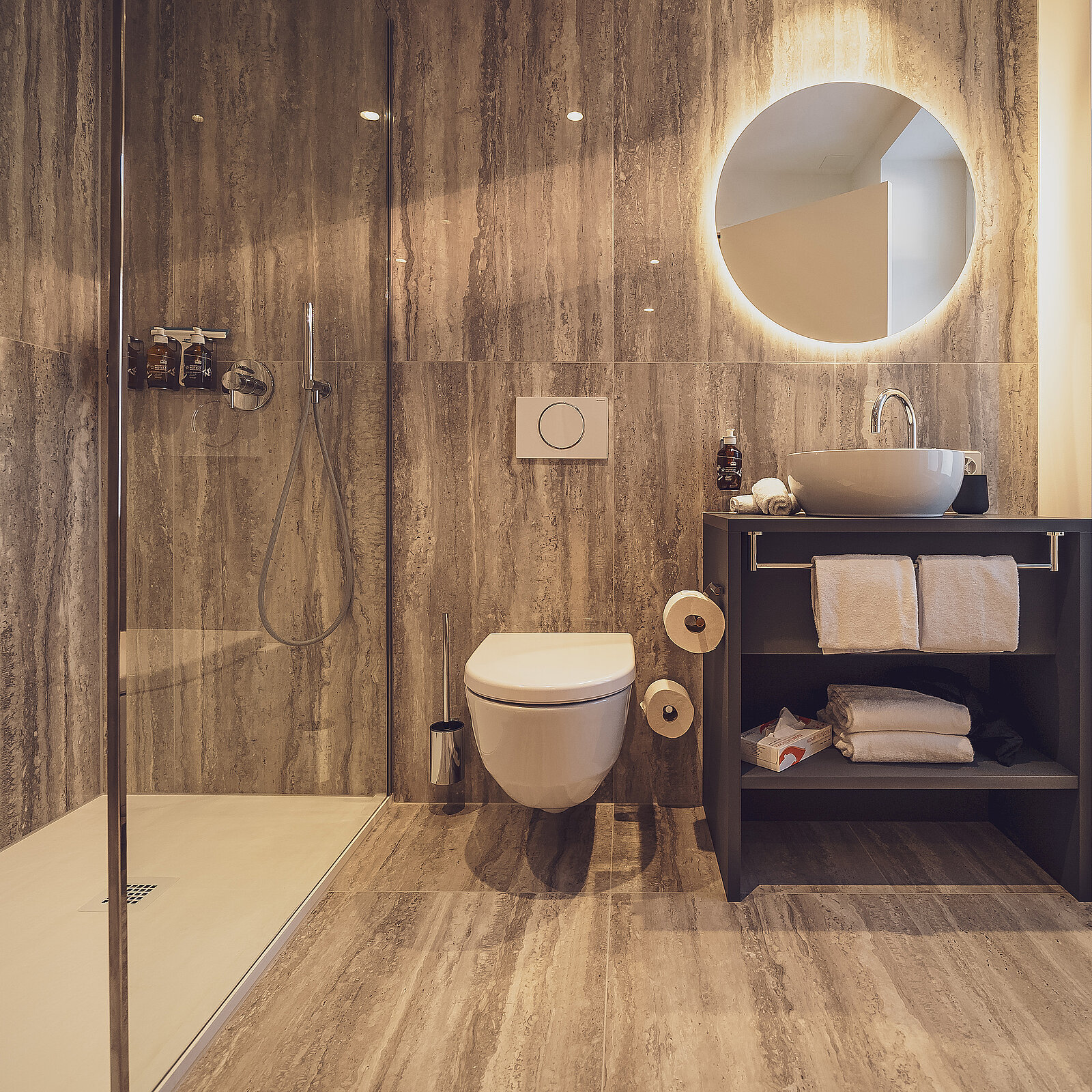 Ein modernes Badezimmer mit einer begehbaren Dusche, einer Toilette und einem Waschtisch mit rundem beleuchtetem Spiegel. Der Waschtisch hat ein rundes Waschbecken und darunter gestapelte Handtücher. Das Design ist minimalistisch und elegant mit warmen Beleuchtungselementen.