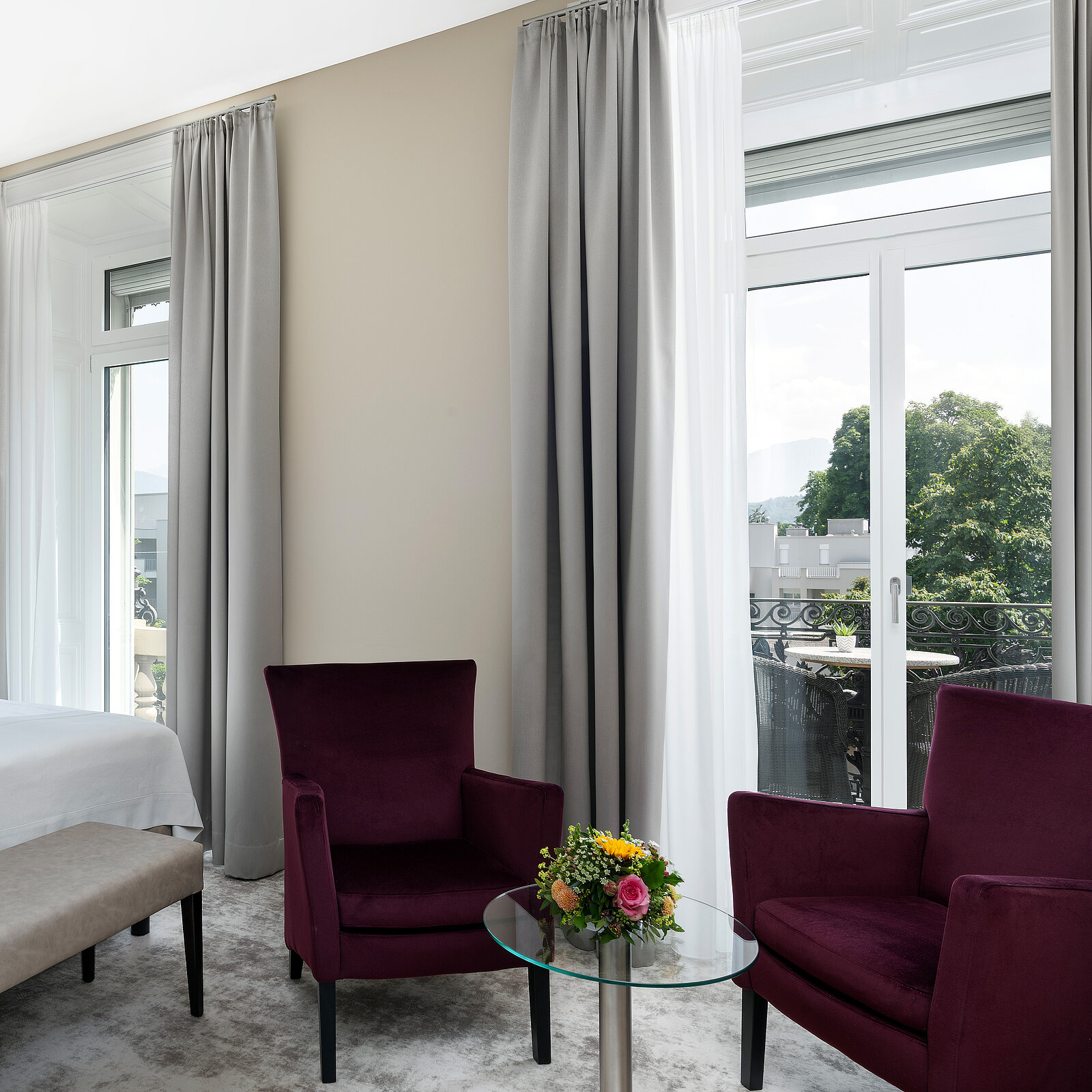 Ein helles Zimmer mit zwei dunkelvioletten Samtsesseln, einem kleinen Glastisch und einem Blumenbouquet darauf. Große Fenster mit weißen und grauen Vorhängen bieten Blick auf einen Balkon und Bäume. Ein Bett und eine Bank sind im Hintergrund sichtbar.