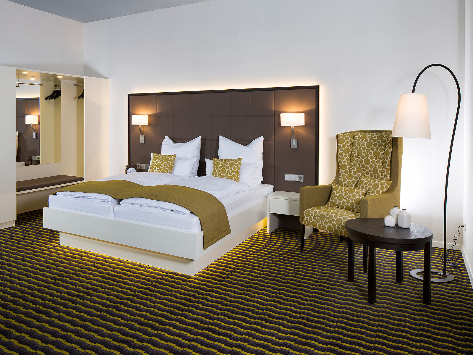 Ein modern gestaltetes Schlafzimmer mit einem Doppelbett, das mit weißen und gelben Textilien ausgestattet ist. Neben dem Bett stehen Nachttische mit Lampen. In der Ecke befindet sich ein Sessel und ein kleiner Tisch. Der Boden ist mit einem gestreiften Teppich ausgelegt.