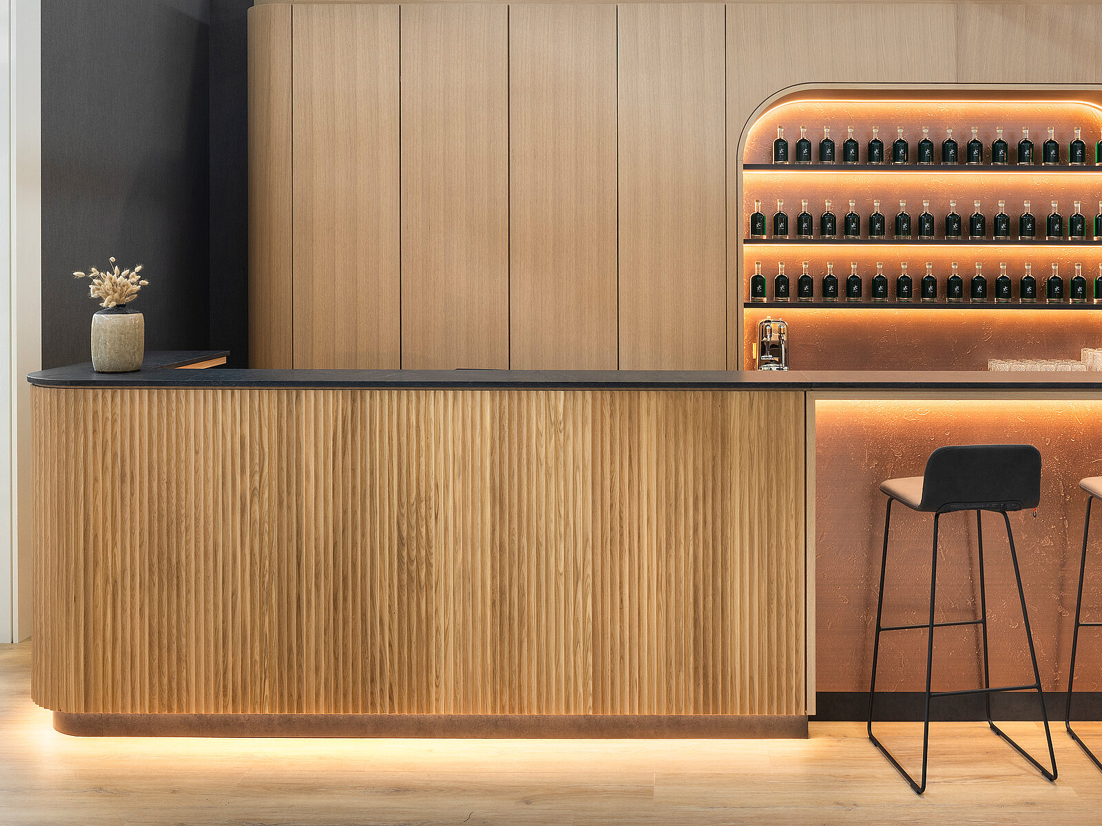 A modern wooden reception counter features a textured surface and is complemented by a sleek black top. Behind it, a softly illuminated shelf displays numerous small dark bottles. Two minimalist black stools are positioned in front of the counter, creating a stylish and welcoming atmosphere.