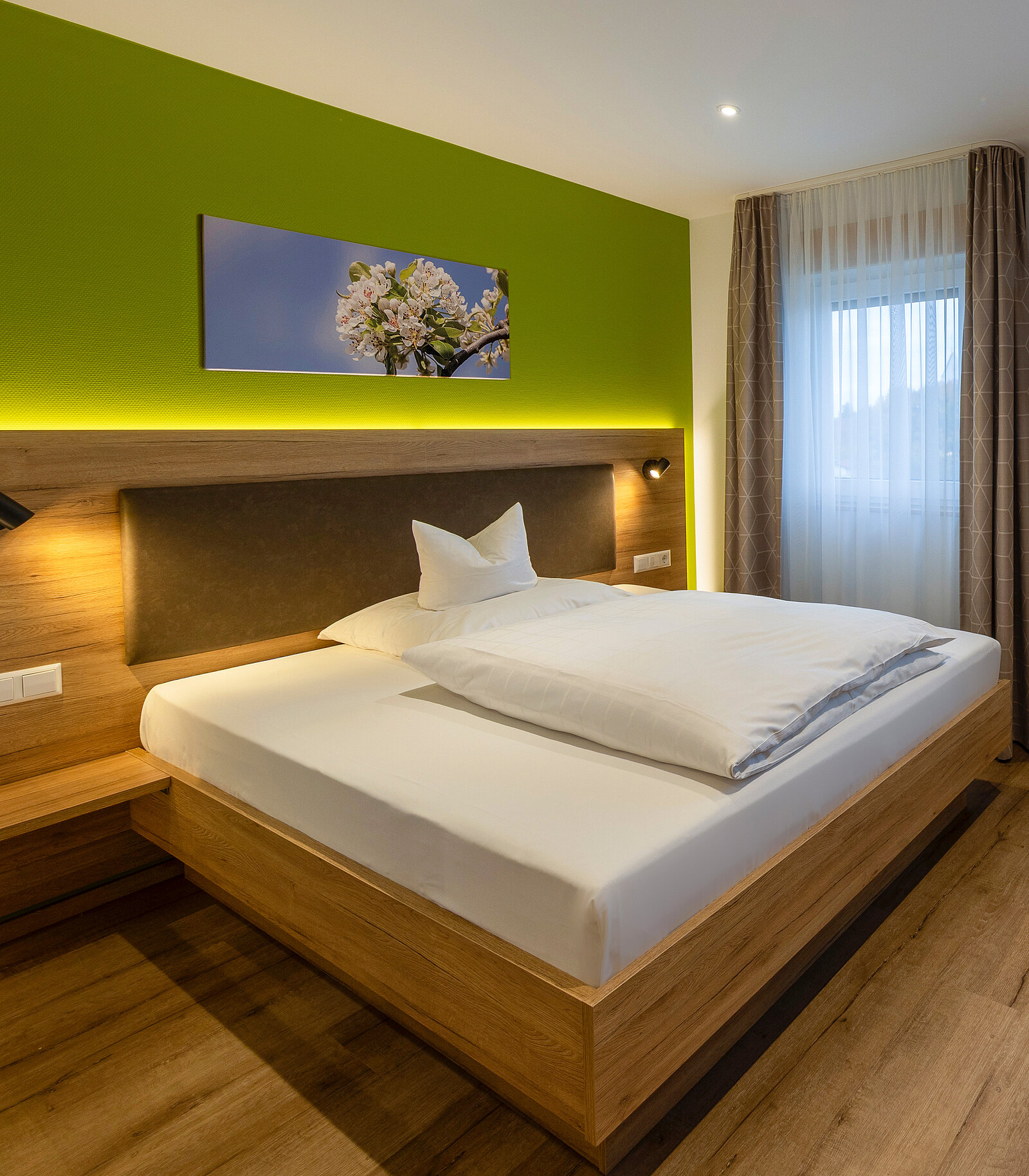 Gemütliches Schlafzimmer mit einem großen Holzbett, weißer Bettwäsche und einem dekorativen Kissen. An der grünen Wand hängt ein Bild mit Blütenmotiv. Ein Fenster mit Vorhängen lässt Tageslicht herein. Dezente Beleuchtung sorgt für eine warme Atmosphäre.