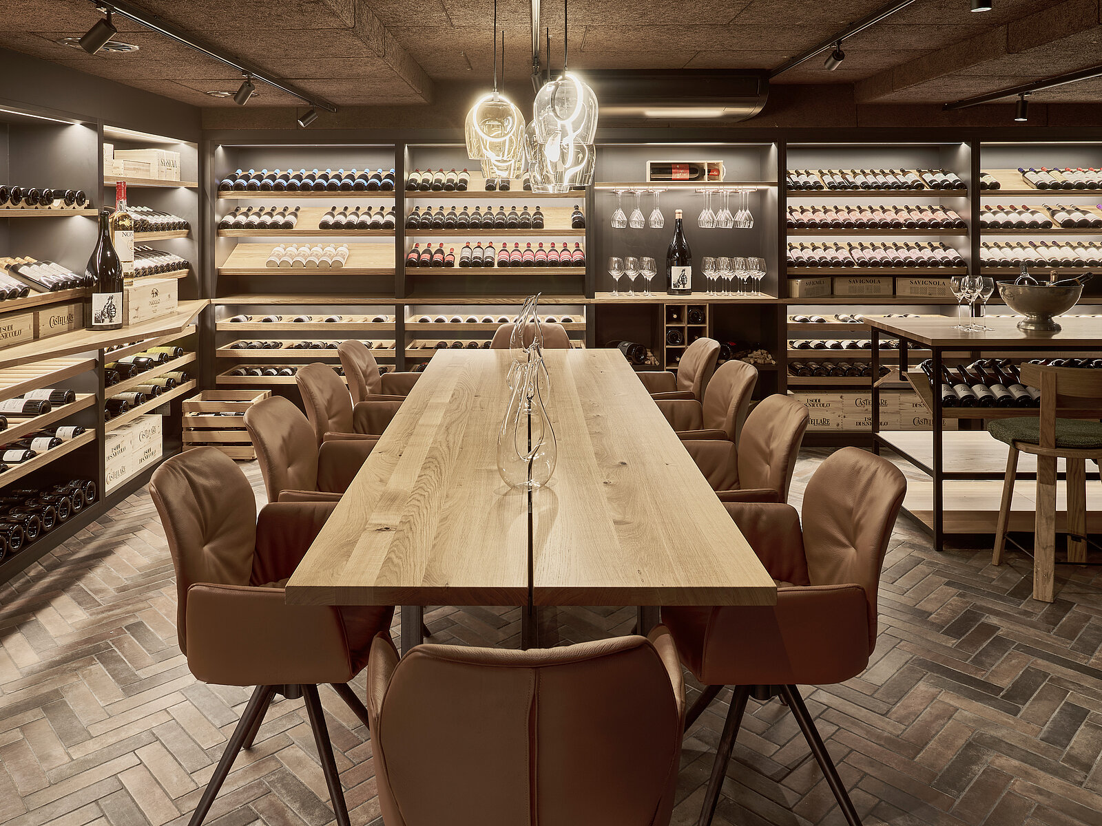 A stylish wine cellar features a long wooden table surrounded by leather chairs. Shelves filled with bottles of wine line the walls, and a modern light fixture hangs above the table, creating an inviting atmosphere for wine appreciation or dining.