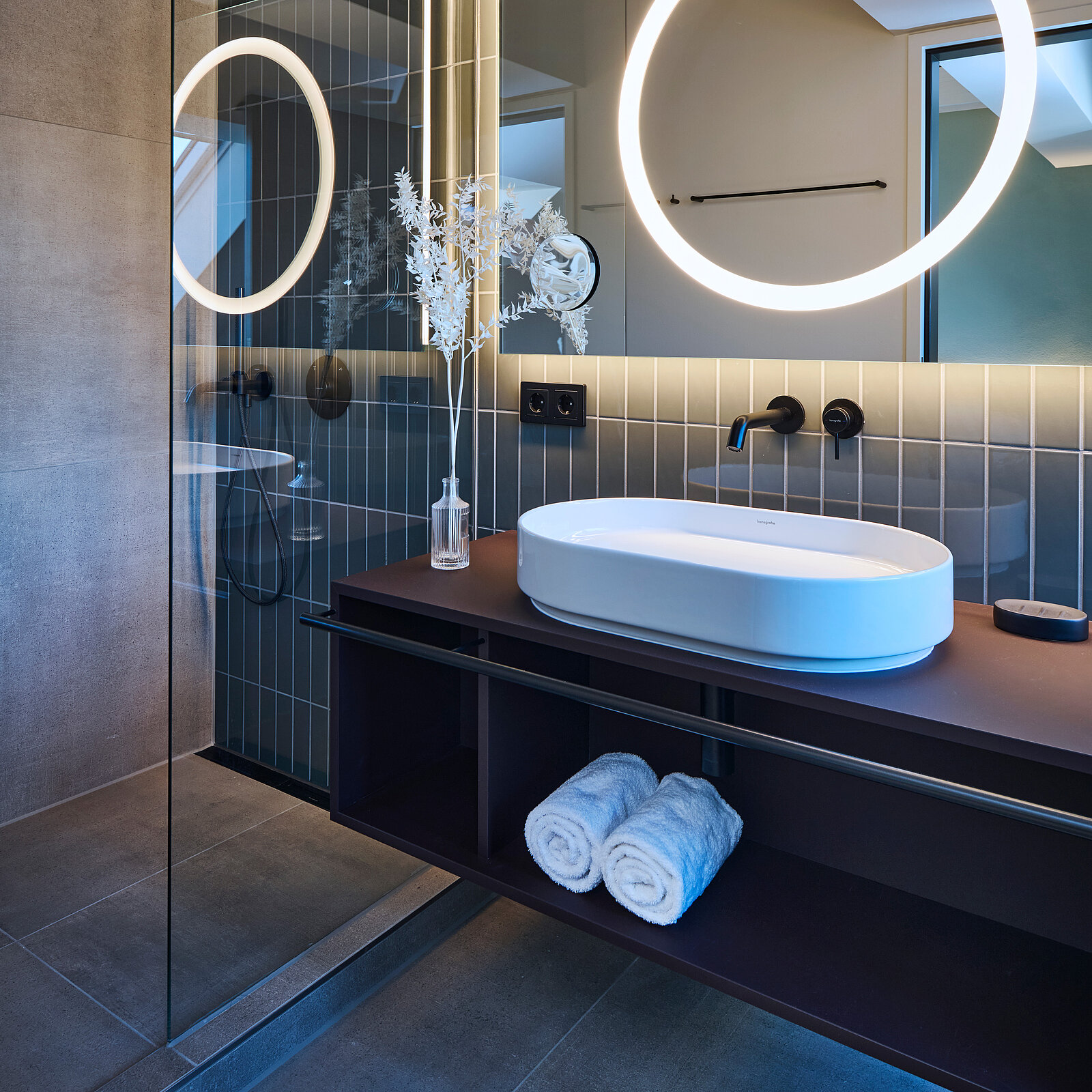 Modernes Badezimmer mit Waschbecken auf dunkler Konsole, rundem beleuchtetem Spiegel und dekorativen Zweigen in einer Vase. Elegante Gestaltung mit dunklen Fliesen, Glasduschabtrennung und gefalteten Handtüchern.