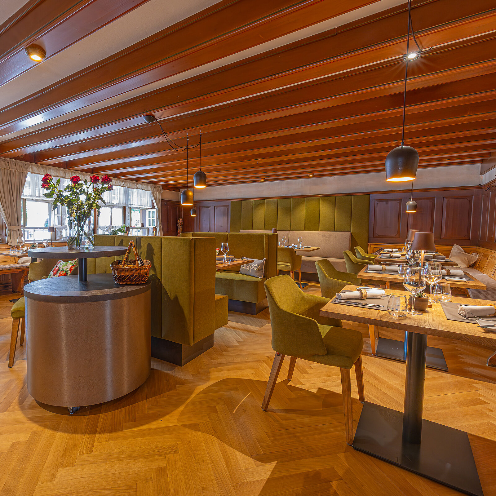 Gemütliches Restaurant mit Holzboden und grünen Polsterbänken. Tische sind stilvoll mit Geschirr und Gläsern gedeckt. Dezente Beleuchtung und ein großer Blumenstrauß ergänzen das Ambiente.