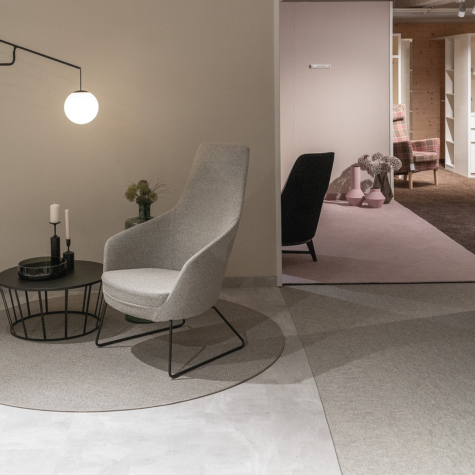 Ein modernes Wohnzimmer mit einem gemütlichen, grauen Stuhl neben einem runden Couchtisch. Auf dem Tisch steht eine Kerze und eine kleine Pflanze. Im Hintergrund ist ein sanft beleuchteter Raum mit Regalen und einem weiteren Stuhl zu sehen.