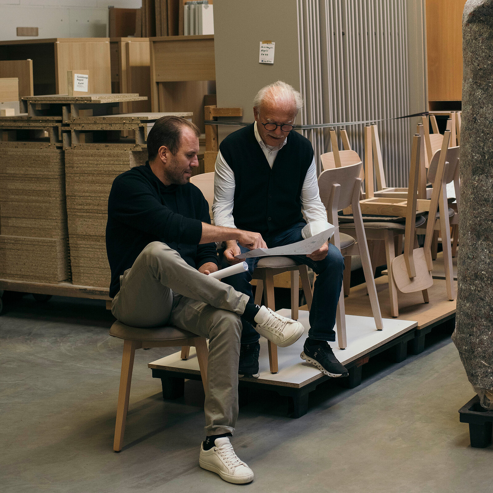 Two men are seated in a workshop discussing plans. One man points at a blueprint while the other examines it. Surrounding them are various pieces of furniture, creating a collaborative and creative atmosphere.
