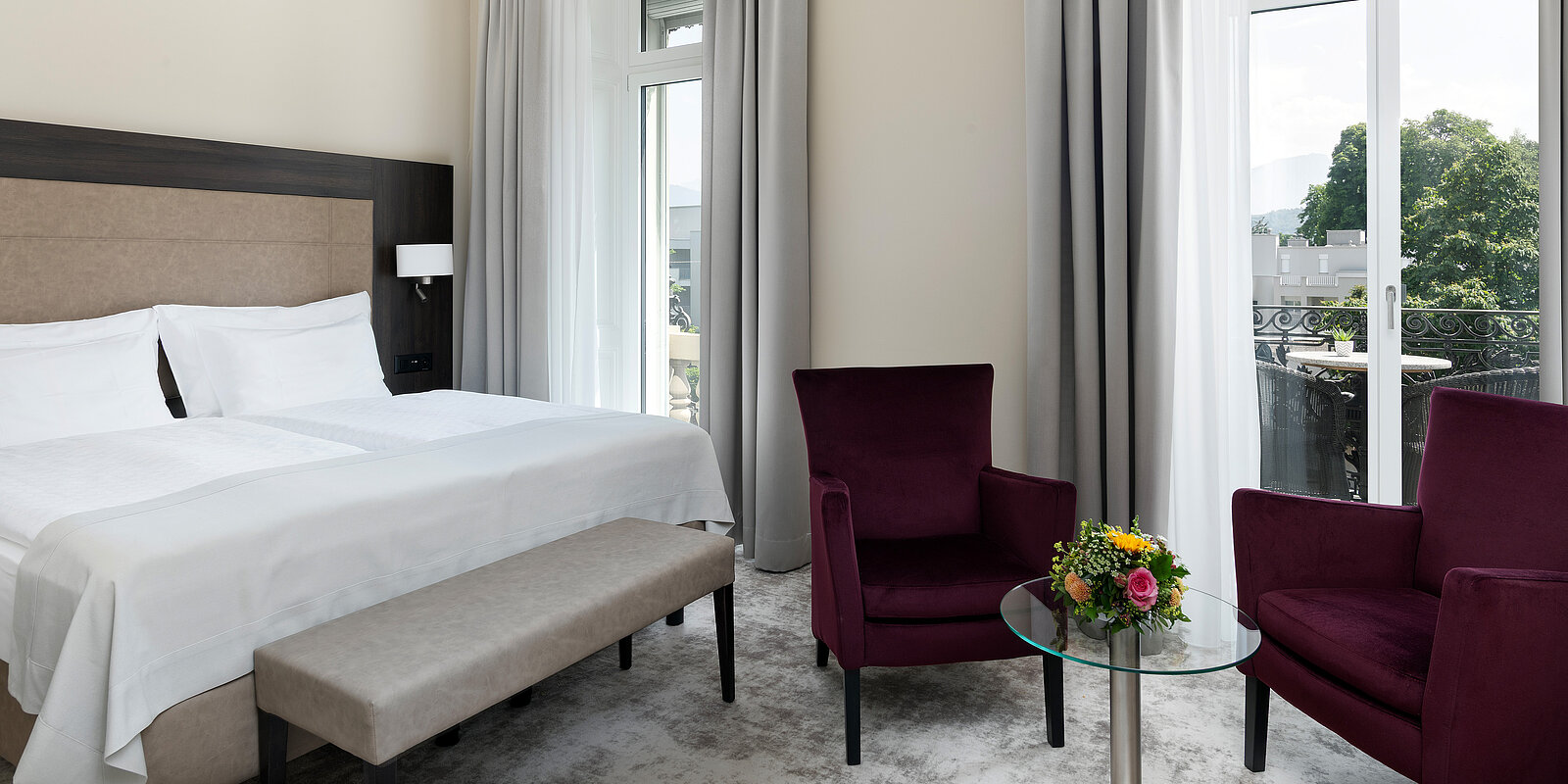 Helles Hotelzimmer mit großem Doppelbett, zwei lila Sessel und einem kleinen Glastisch mit Blumenstrauß. Große Fenster mit weißen Vorhängen bieten Blick nach draußen. Elegantes und modernes Design sorgt für eine einladende Atmosphäre.