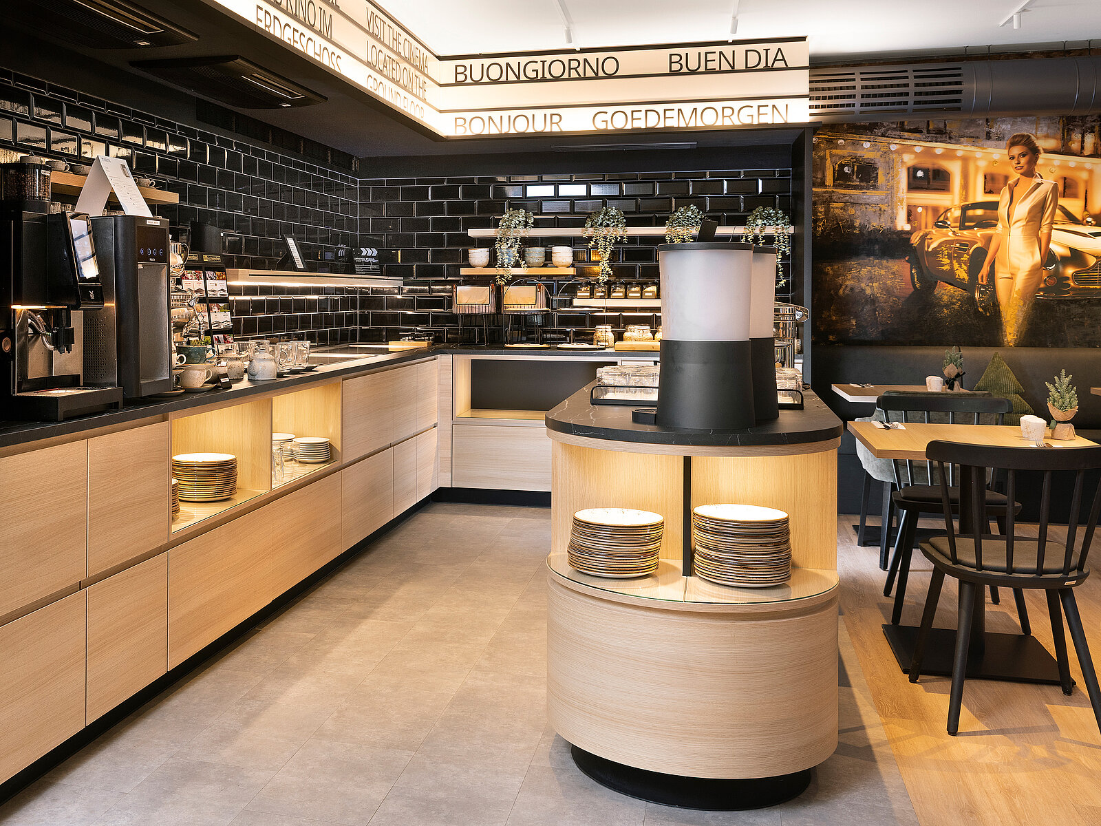 A modern café interior featuring warm wood tones and sleek black accents. The space includes a coffee machine, neatly arranged plates on a circular counter, and decorative plants. On the walls, welcoming phrases in multiple languages greet customers, creating an inviting atmosphere.