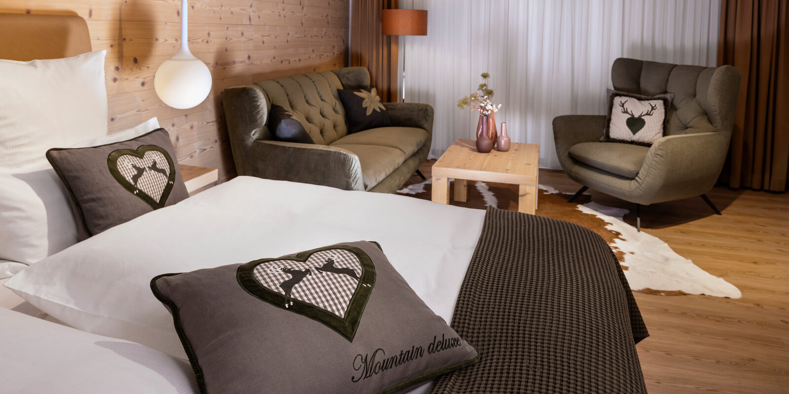 zieflekoch musterzimmer zulux, gemütliches Zimmer mit einem Doppelbett, dekorativen Kissen, einem Sofa, einem Sessel, Holzwänden und einem kleinen Tisch mit Vasen, warme Beleuchtung und Holzfußboden