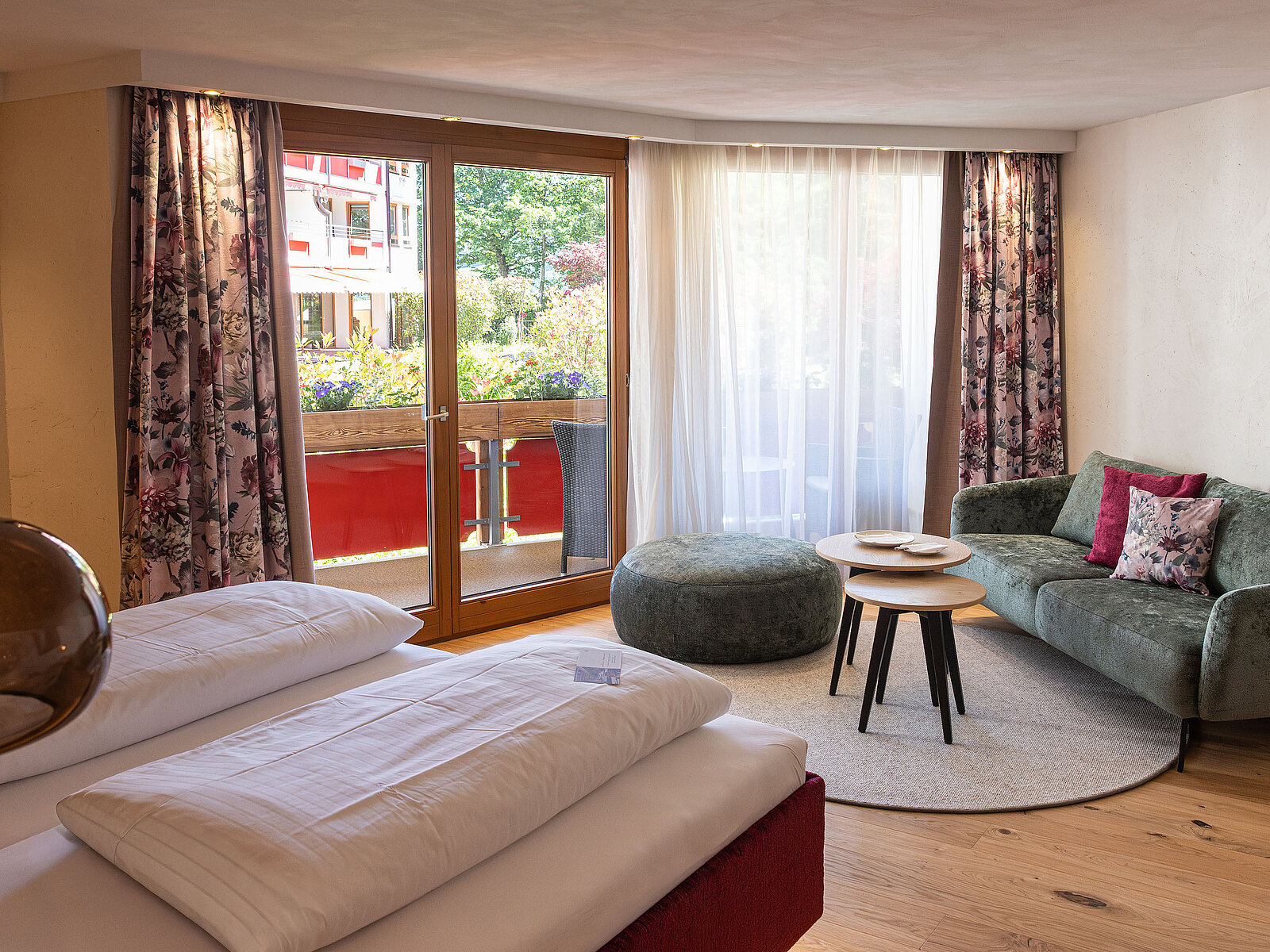 Gemütliches Hotelzimmer mit Doppelbett, kleinem Sofa und Couchtisch. Große Fensterfront mit Zugang zu einem blumengeschmückten Balkon. Helle, freundliche Atmosphäre mit stilvollen Dekorationen.