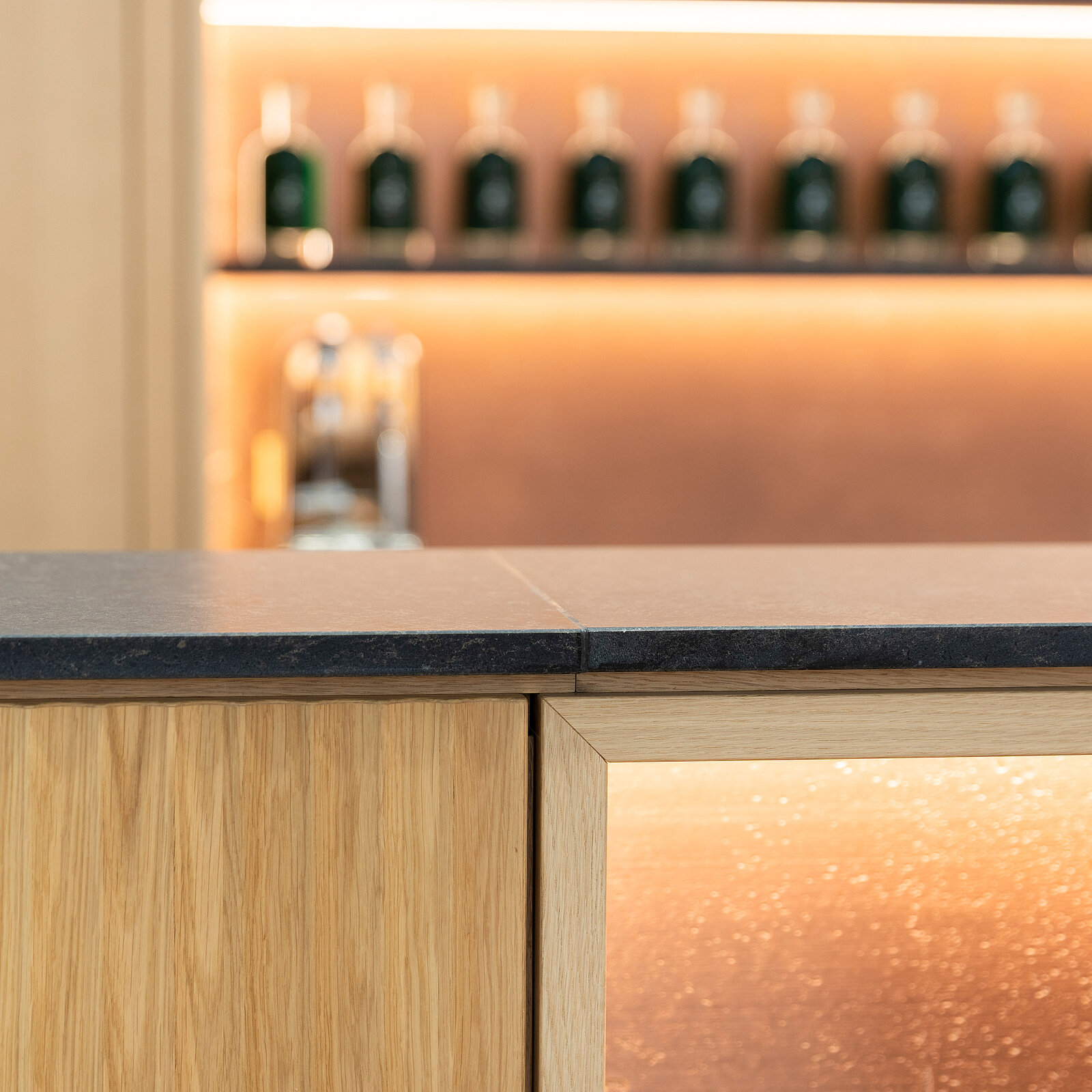 A sleek bar counter made of dark stone features warm backlighting. Behind the counter, a shelf displays several dark glass bottles, creating a modern and inviting atmosphere. The design combines natural wood elements with contemporary lighting.