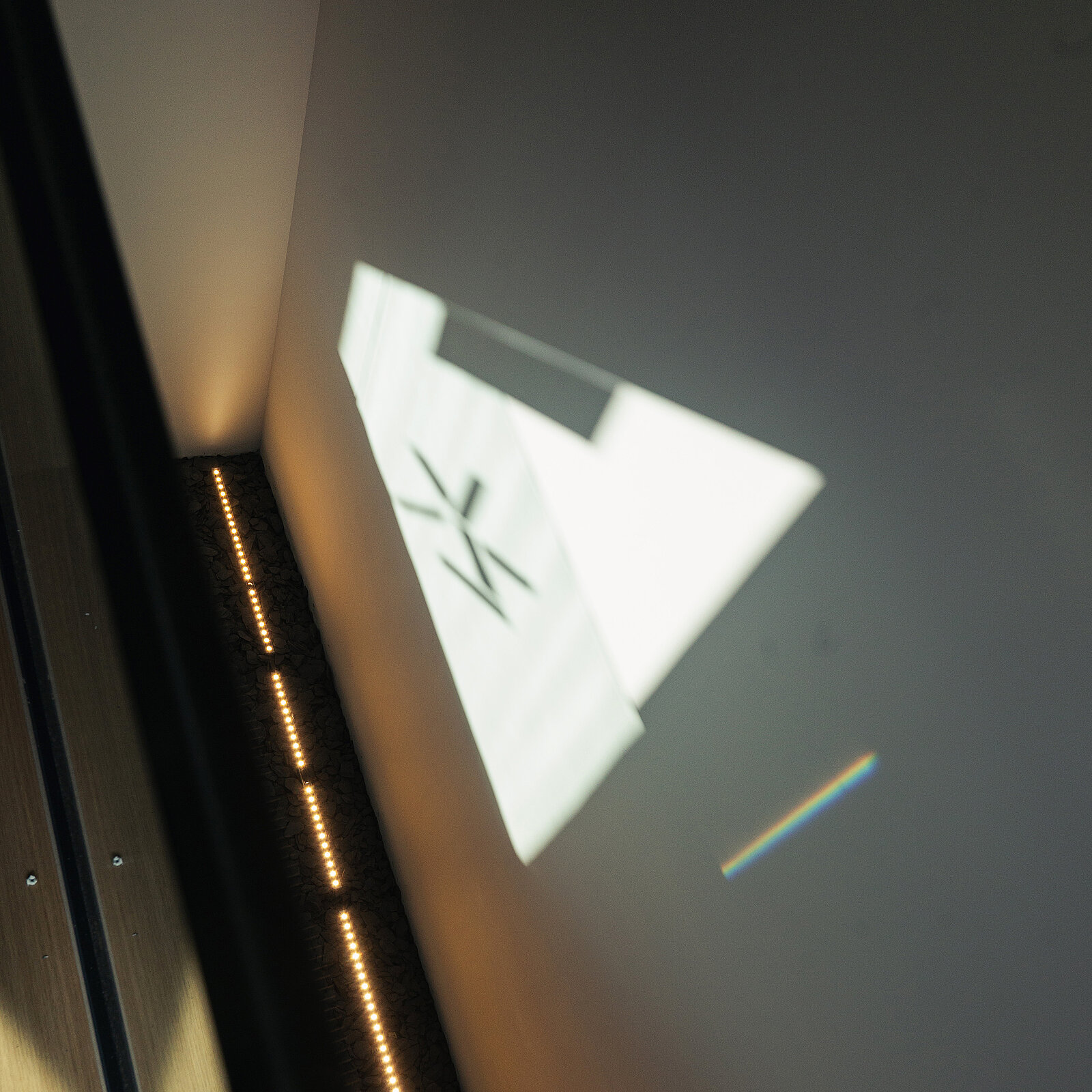 Eine graue Wand in einem Aufzug mit einem projizierten, weißen Symbol, das ein schlichtes "zk" darstellt. Am Boden sind weiche, warmweiße Lichtleisten sichtbar. Das Bild vermittelt ein Gefühl von Modernität und minimalistischem Design.