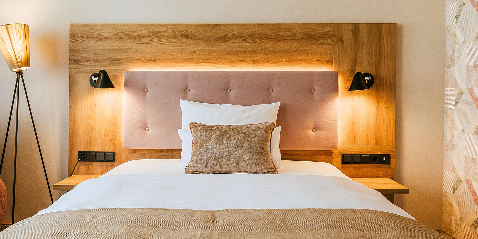 Das Bild zeigt ein stilvolles Schlafzimmer mit einem modernen Bett. Der Kopfteil aus Holz hat eine gepolsterte, helle Fläche. Rechts und links vom Bett sind Wandleuchten angebracht. Auf dem Bett liegt ein dekoratives Kissen und die Bettwäsche ist schlicht und hell.