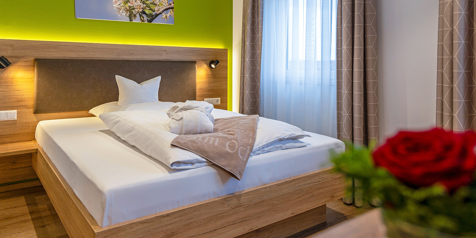 Modern eingerichtetes Hotelzimmer mit einem großen Bett, auf dem zwei weiße Kissen und ein gefaltetes Handtuch liegen. Im Hintergrund befindet sich ein beleuchtetes Kopfteil, während im Vordergrund eine rote Rose in einer Vase auf einem Tisch steht.