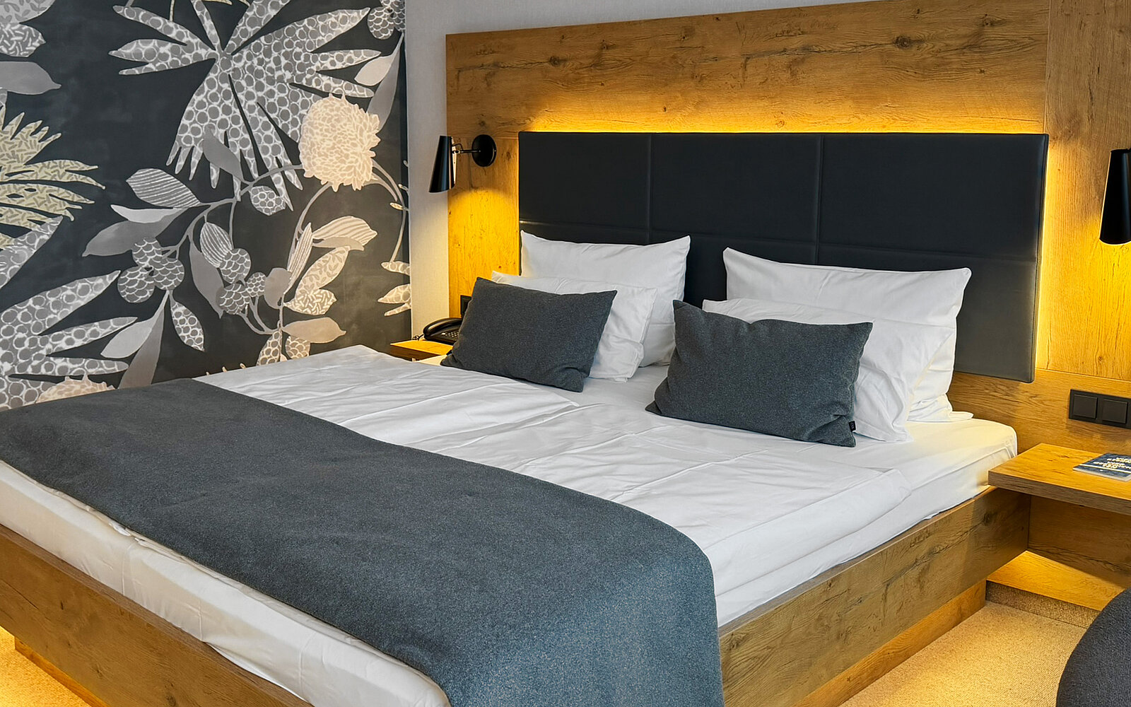 Das Bild zeigt ein modernes Schlafzimmer mit einem großen Doppelbett, das mit weißen Bettlaken und grauen Kissen dekoriert ist. Hinter dem Bett befindet sich eine beleuchtete Wand mit einem dezenten Muster von Pflanzen. Eine Holzkommode steht neben dem Bett.