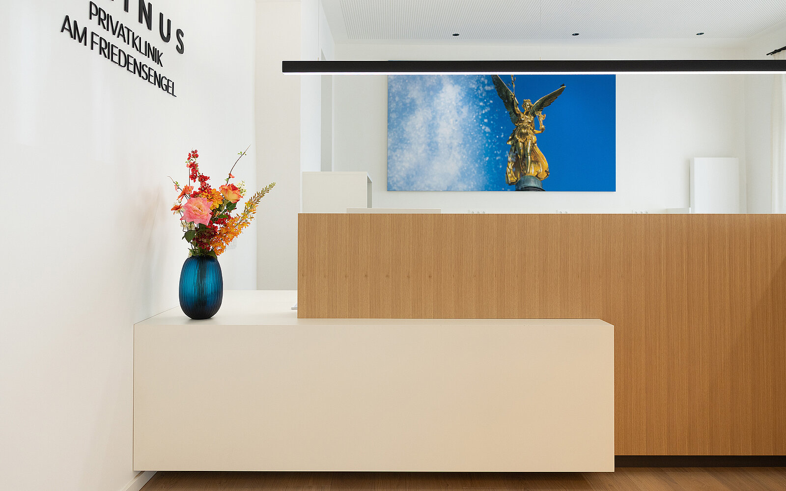 Ein moderner Empfangsbereich mit einem weißen Tresen und hölzernen Elementen. Ein blauer Vase mit bunten Blumen steht auf der Theke. An der Wand hängt ein großes Bild einer goldenen Statue vor blauem Himmel.