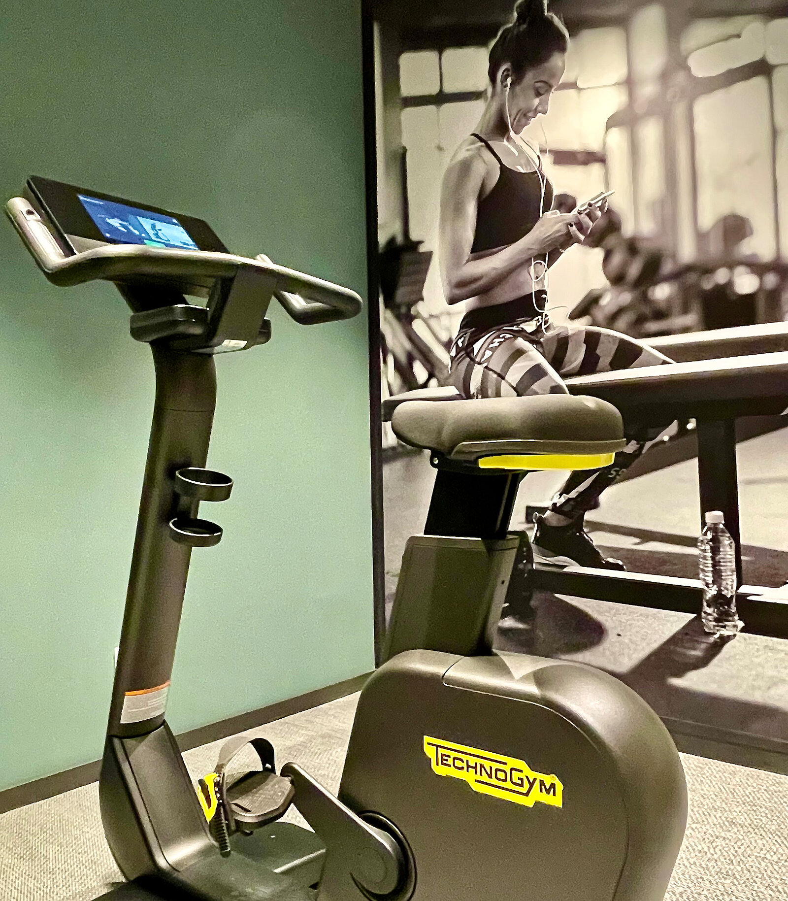 Ein modernes Ergometer mit einem digitalen Bildschirm steht vor einer Wand mit einem Bild einer fitten Person, die beim Training auf ihr Handy schaut. Die Umgebung ist sportlich und motivierend gestaltet.