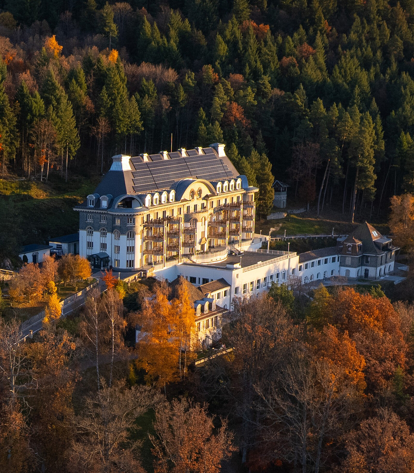 Ein großes, elegantes Hotelgebäude steht im Herbstwald. Das Gebäude hat eine auffällige Architektur mit zahlreichen Fenstern und einem charakteristischen Dach. Rundherum sind Bäume mit buntem Laub in verschiedenen Herbstfarben.