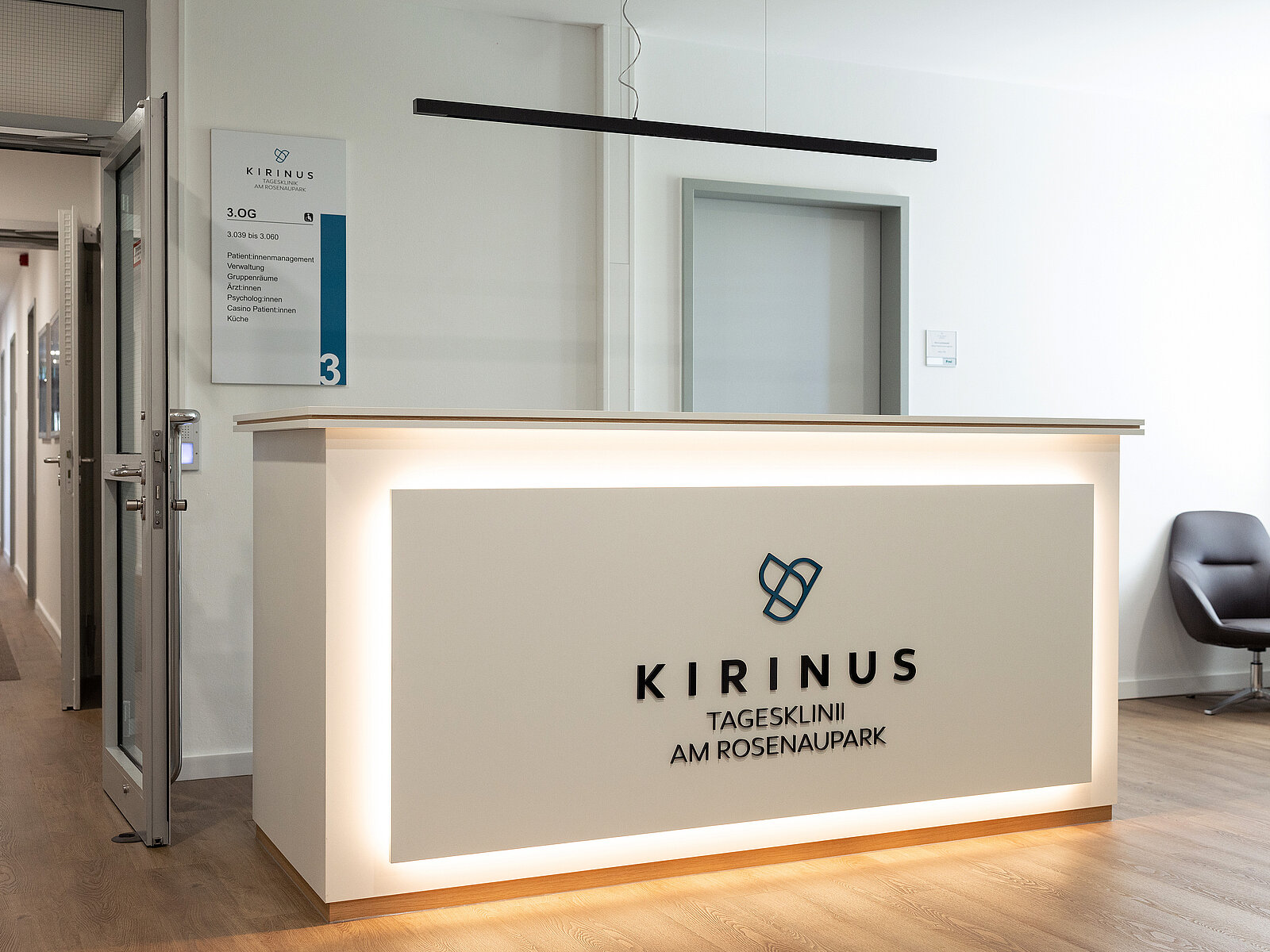 Die Rezeption der Kirinus Tagesklinik am Rosenaupark. Der Empfangstresen ist modern gestaltet, mit dem Logo und Namen der Klinik darauf. Daneben befindet sich eine gepolsterte schwarze Sitzgelegenheit. Ein Informationsschild hängt an der Wand neben einer Tür mit der Nummer 300.