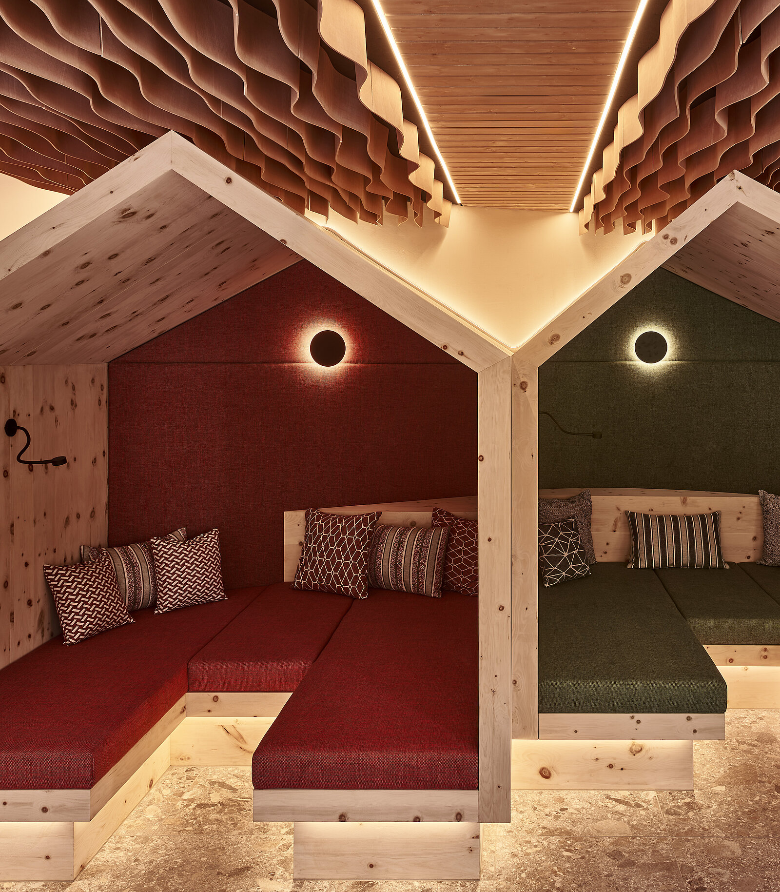 The image features a cozy interior design with two stylized, wooden "house" structures. Each house has a padded seating area, one with red fabric and the other in green. Soft ambient lighting complements a warm, inviting atmosphere, creating a comfortable space for relaxation.