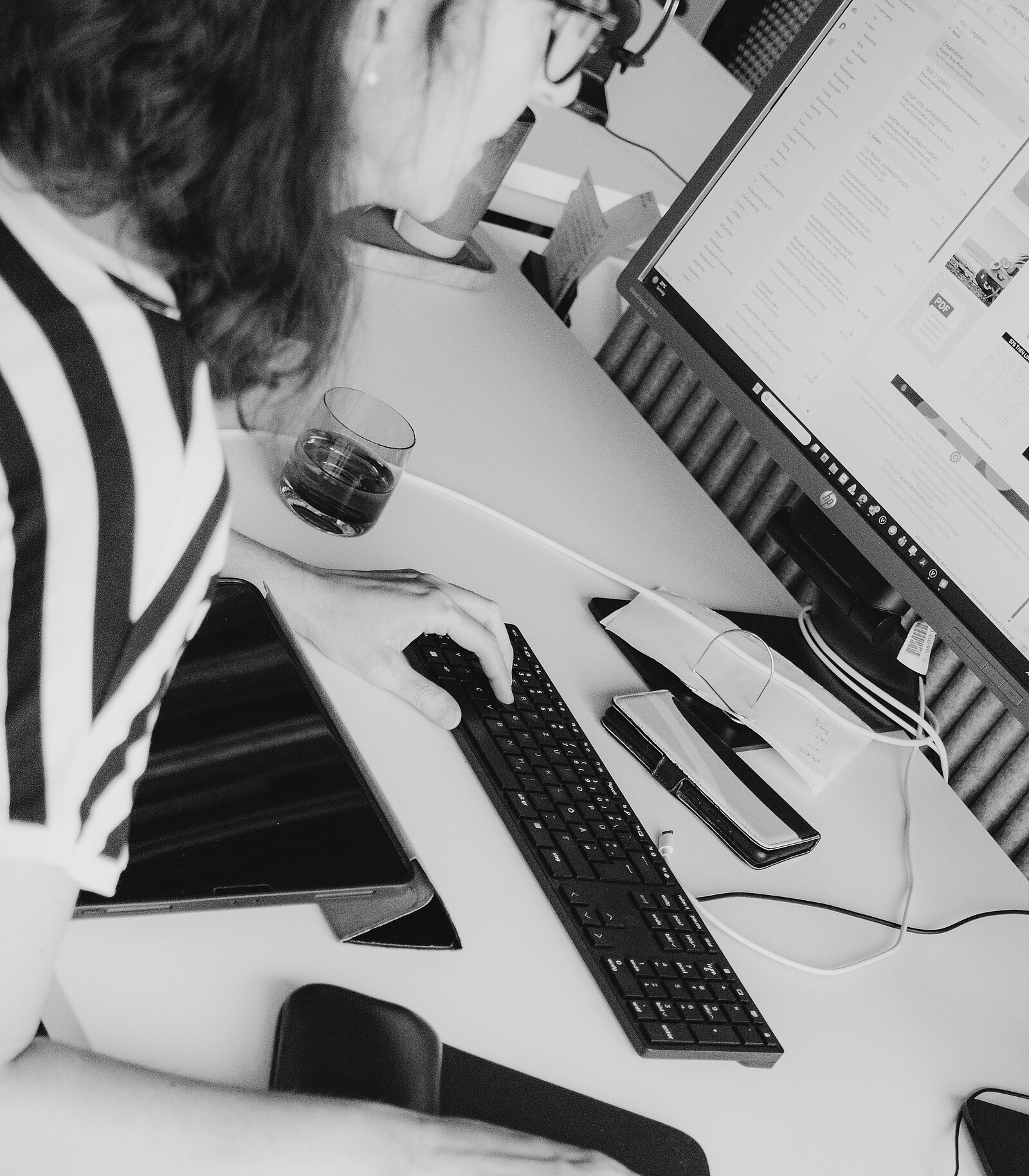 Eine Person sitzt an einem Schreibtisch und arbeitet am Computer. Der Bildschirm zeigt eine Webseite, und neben der Tastatur liegt ein Tablet. Ein Glas Wasser steht in der Nähe.