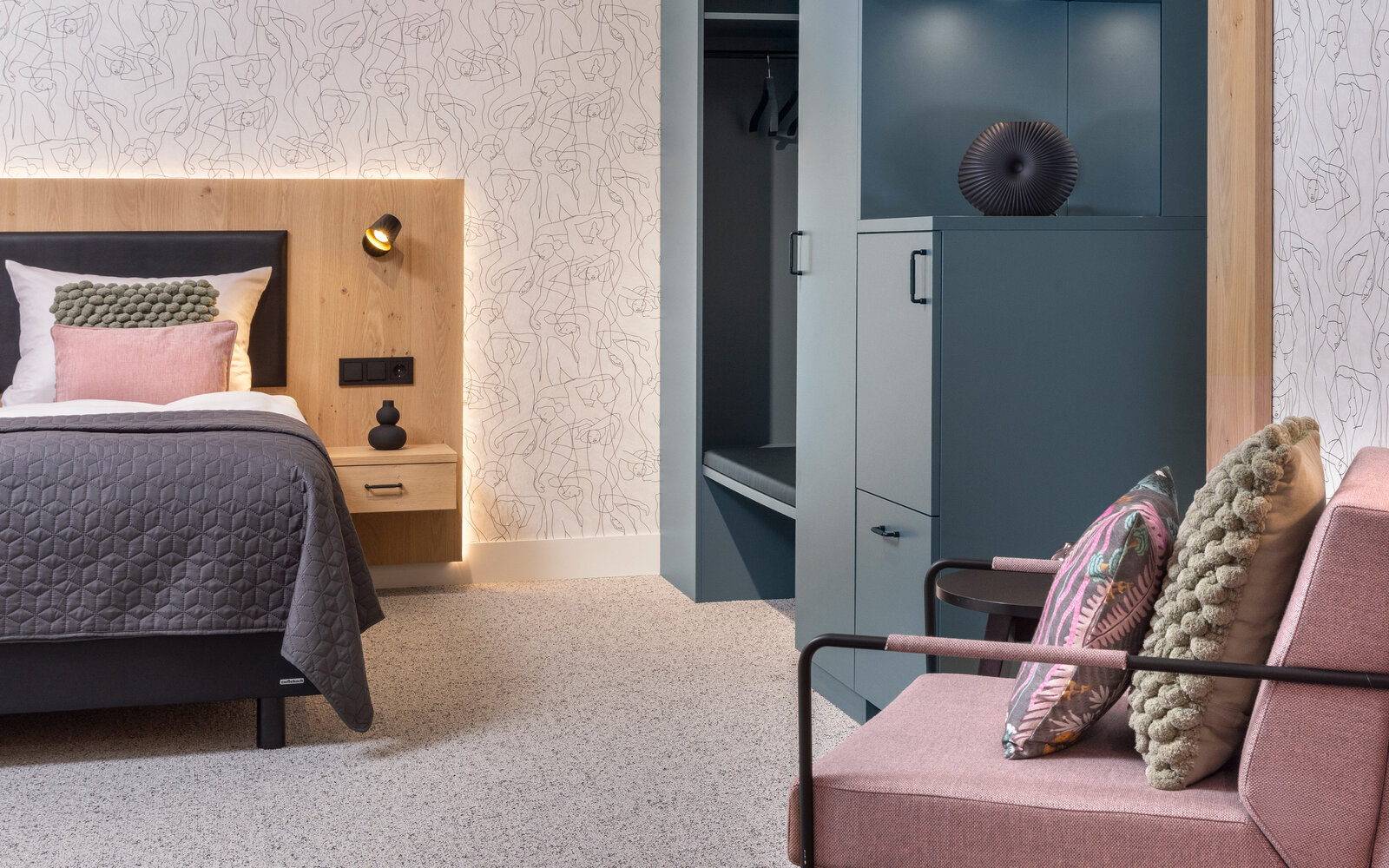 zieflekoch musterzimmer zenit, modernes Schlafzimmer mit einem Boxspring-Doppelbett mit Rückwand in Holzoptik, einem rosafarbenen Sessel, einem Schrank in gedecktem Grün und einer abstrakten Motivtapete, der Teppichboden und ein dunkler Teppich verleihen dem Raum Wärme.