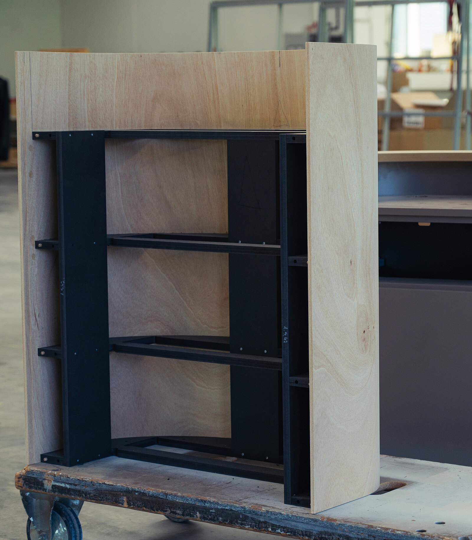 Ein unvollständiger Möbelstück, bestehend aus einem hellen Holzrahmen mit offenen Regalböden aus schwarzem Metall. Die Konstruktion steht auf einem Rollwagen und ist in einer Werkstattumgebung platziert.