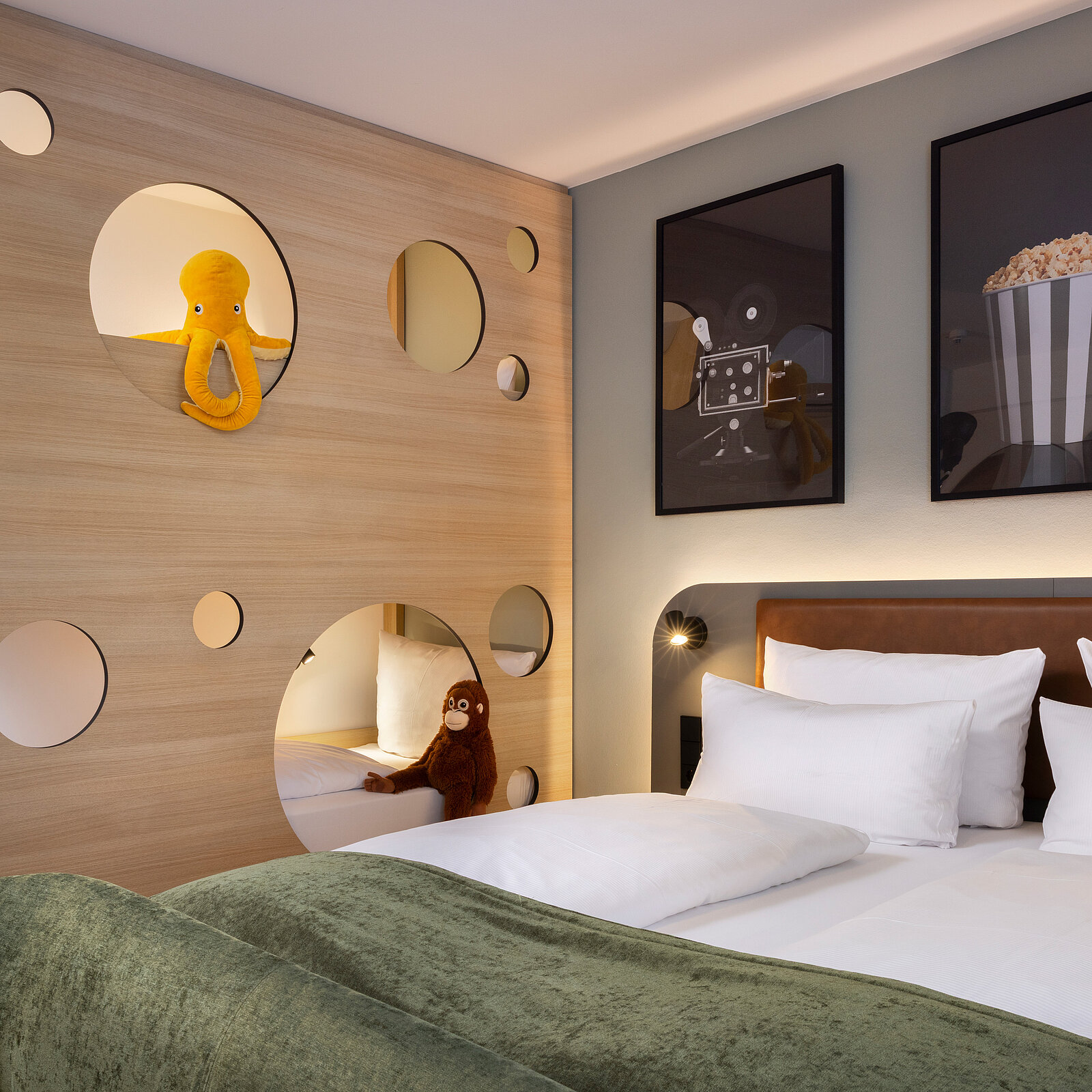 A cozy bedroom features a green bedspread, white pillows, and playful wall decorations. On the left, a wooden partition with circular cutouts shows a yellow octopus and a brown monkey plush toy. Two framed artworks hang above a minimalistic bedside table, enhancing the room's inviting atmosphere.