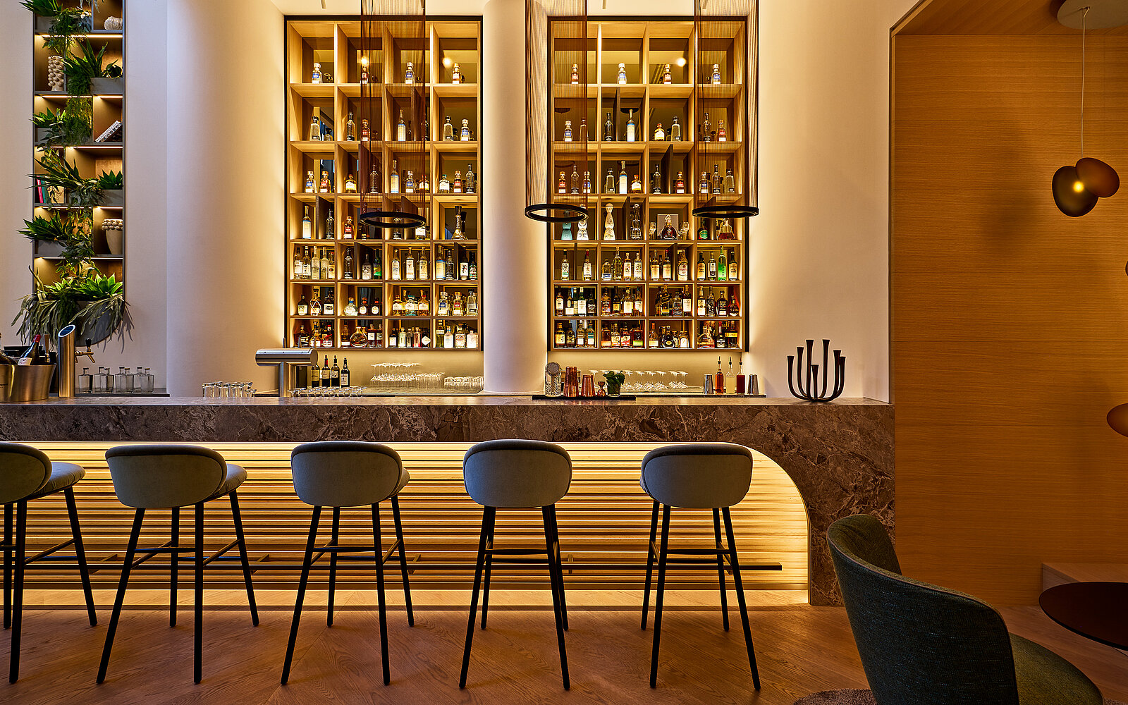 Eine elegant gestaltete Bar mit fünf hohen Barhockern vor einem langen Tresen. Dahinter befinden sich zwei große, beleuchtete Regale, die mit diversen Flaschen bestückt sind. Die Atmosphäre ist warm und einladend durch gedämpfte Beleuchtung und Holzakzente.