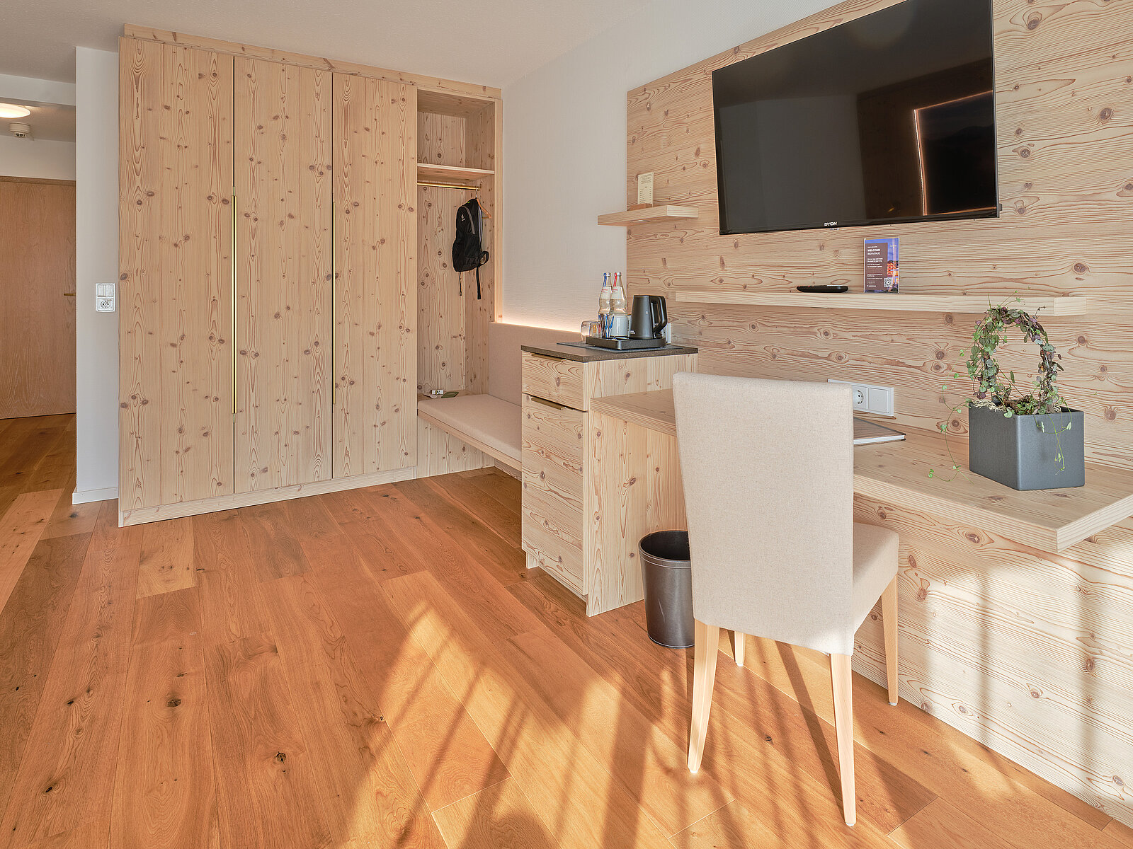 Ein modernes Hotelzimmer mit Holzmöbeln: Kleiderschrank links, Schreibtisch mit Stuhl rechts, darüber ein an der Wand montierter Flachbildfernseher. Der Boden besteht aus Holz. Dezente Beleuchtung sorgt für eine warme Atmosphäre. Ein Regal auf dem Schreibtisch enthält Getränke und eine Pflanze.