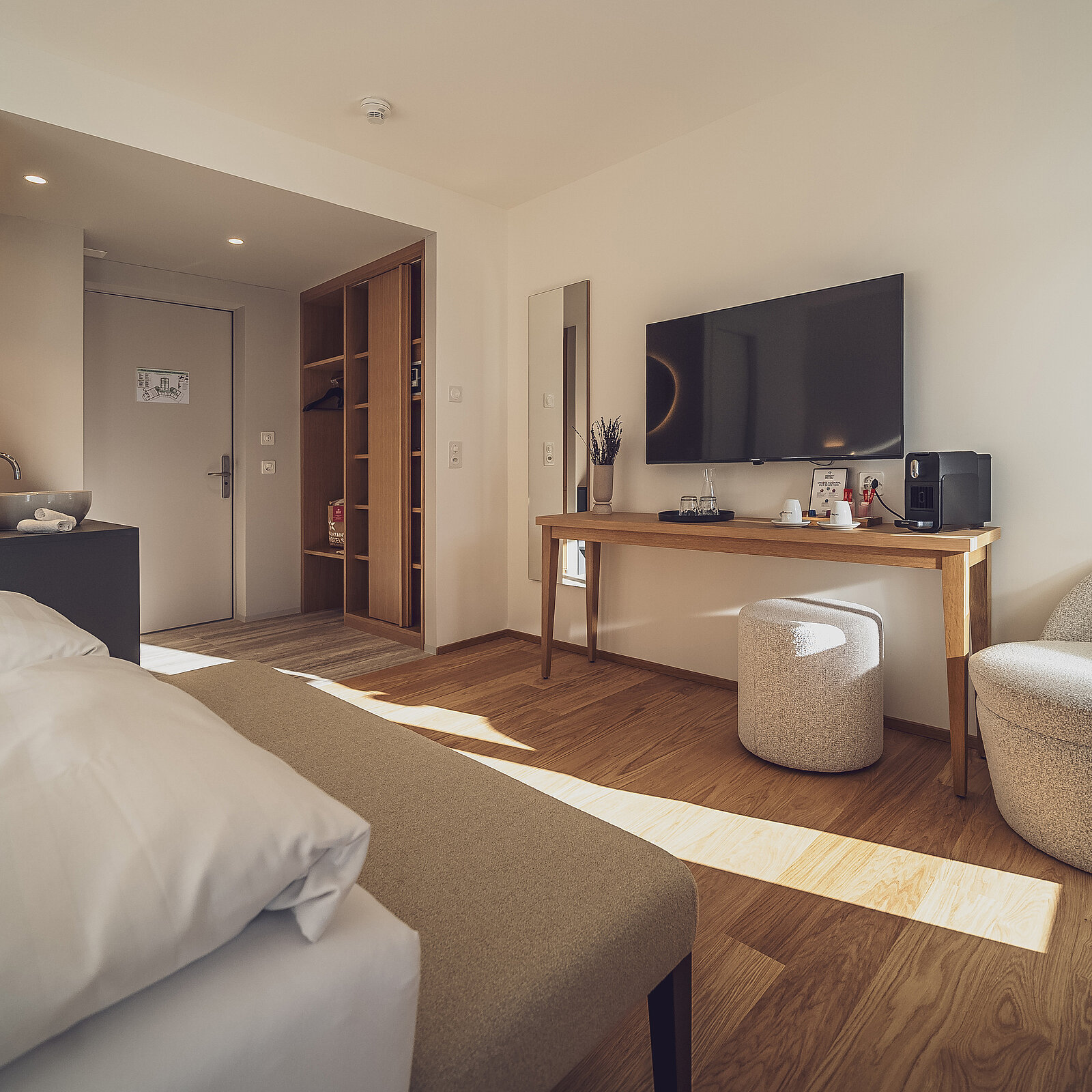 Ein modernes Hotelzimmer mit Doppelbett, Flachbildfernseher an der Wand, einem Holzschreibtisch und einem runden Hocker. Ein großes Fenster lässt viel Tageslicht herein, was den Raum hell und einladend wirken lässt. Der Boden ist aus Holz und es gibt einen offenen Kleiderschrank.