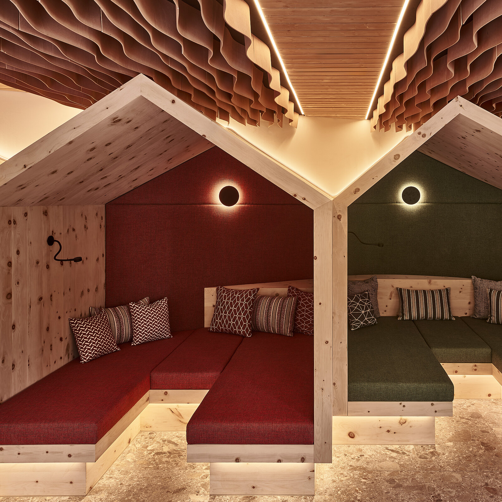 The image features two cozy seating areas designed to resemble small wooden houses. Each area has cushions and ambient lighting, with one side in red and the other in green. The overall atmosphere is warm and inviting, perfect for relaxation or informal gatherings.