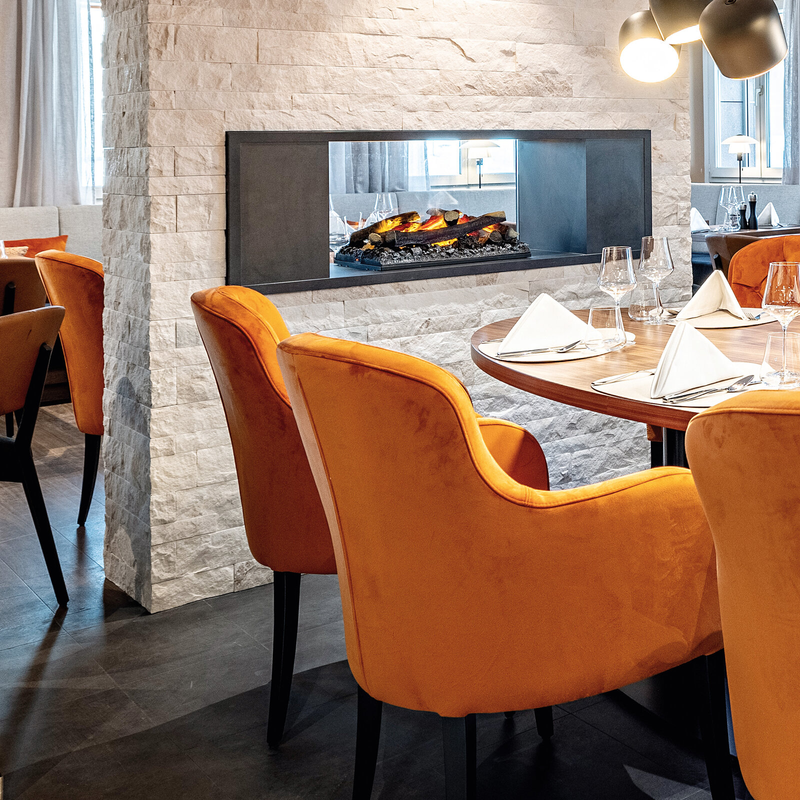 Zieflekoch hotel piz badus Andermatt Schweiz Restaurant Frühstücksraum nussbaum wandbild berge steinwand kamin nussbaum lamellen polstersessel gedeckter Tisch Hängeleuchten winter
