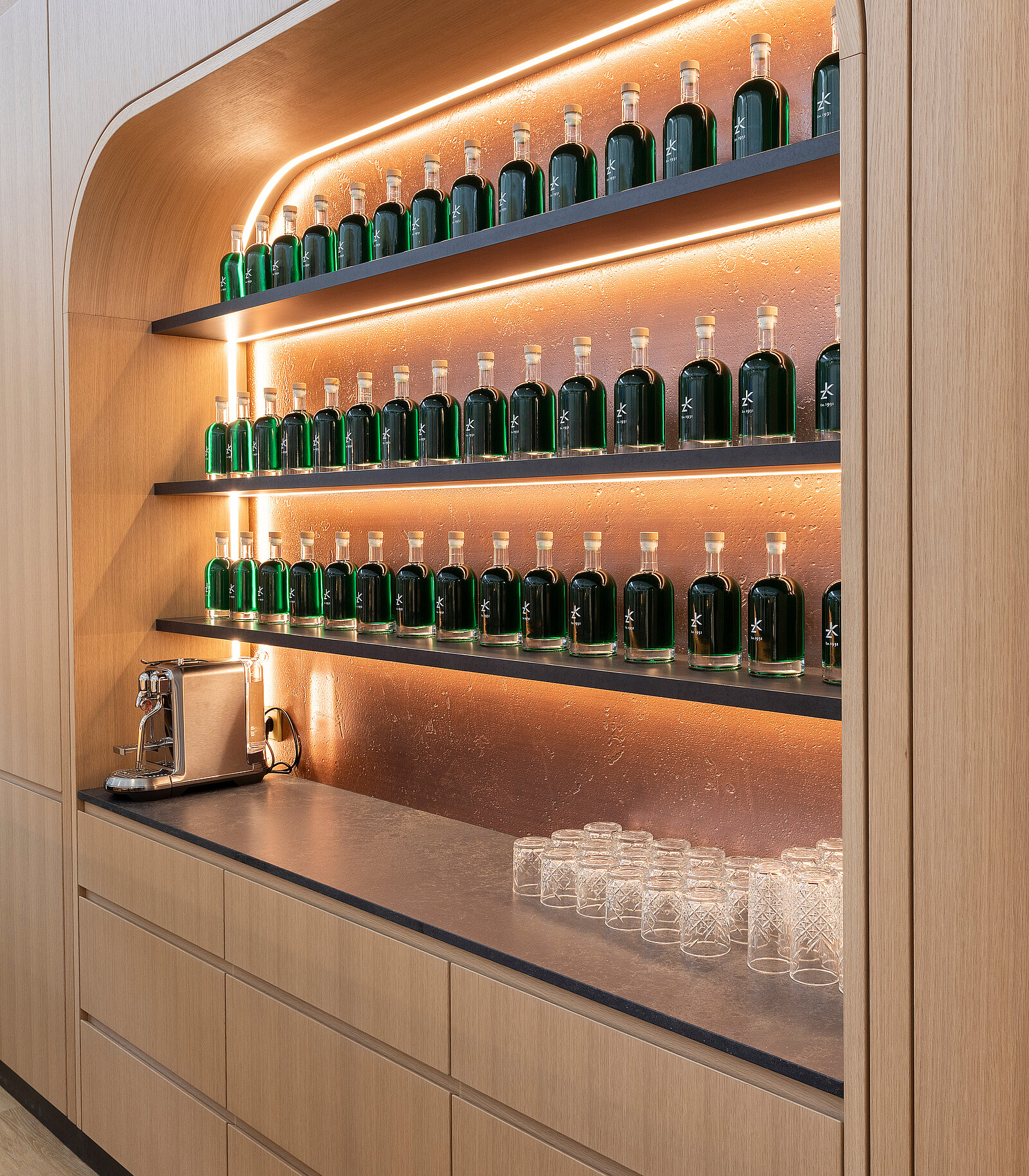A wooden shelving unit displays multiple green glass bottles on two levels, illuminated by soft lighting. Below, there is a countertop with an elegant drink dispenser and several clear glasses arranged neatly. The overall design is modern and minimalist, emphasizing the arrangement of the bottles.