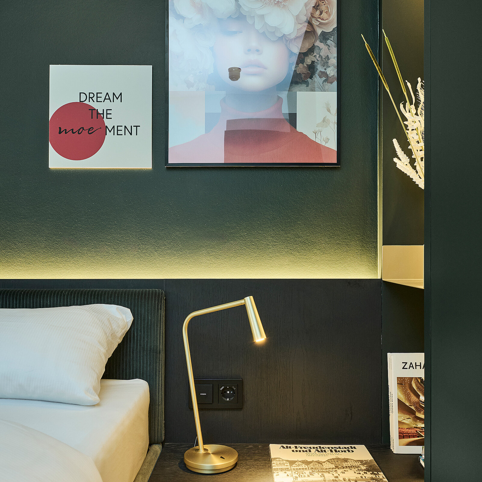 Ein stilvoll eingerichtetes Schlafzimmer mit einem Bett, einer goldfarbenen Leseleuchte und modernen Wandbildern. Ein Bild zeigt eine Frau mit Blumen auf dem Kopf, während ein weiteres Bild die Worte „Dream the Moment“ enthält. Bequeme, minimalistische Atmosphäre.