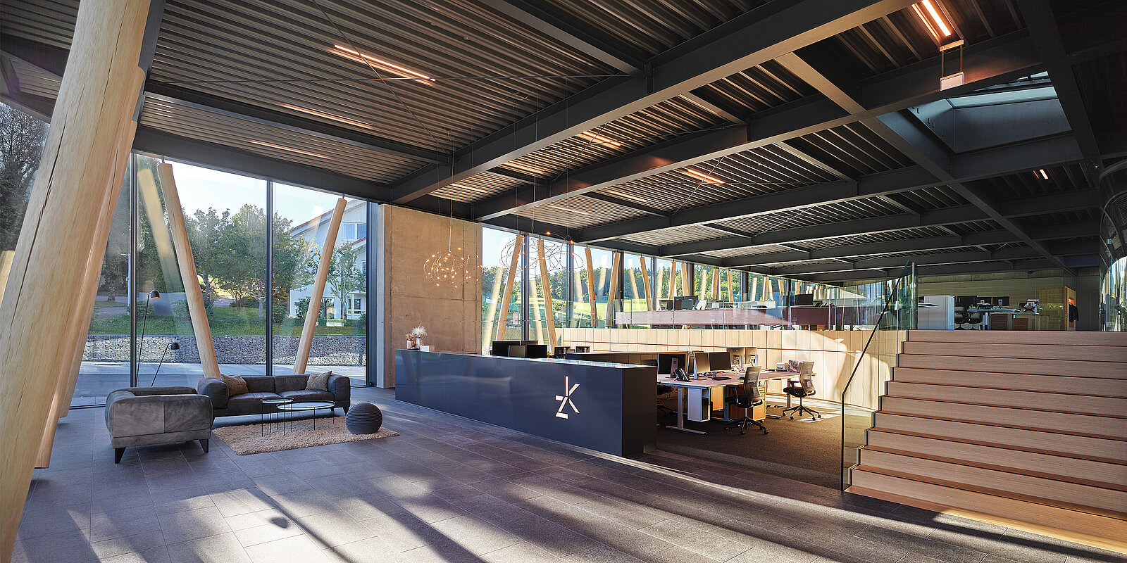 Eine moderne Büroeinrichtung zeichnet sich durch offene Räume mit großen Glasfenstern aus, die Tageslicht hereinlassen. Das Design umfasst Holzbalken und eine Treppe, die zu einer unteren Ebene mit Schreibtischen und Stühlen führt. Ein gemütlicher Sitzbereich mit einem Sofa und einem runden Ottoman schafft eine einladende Atmosphäre.