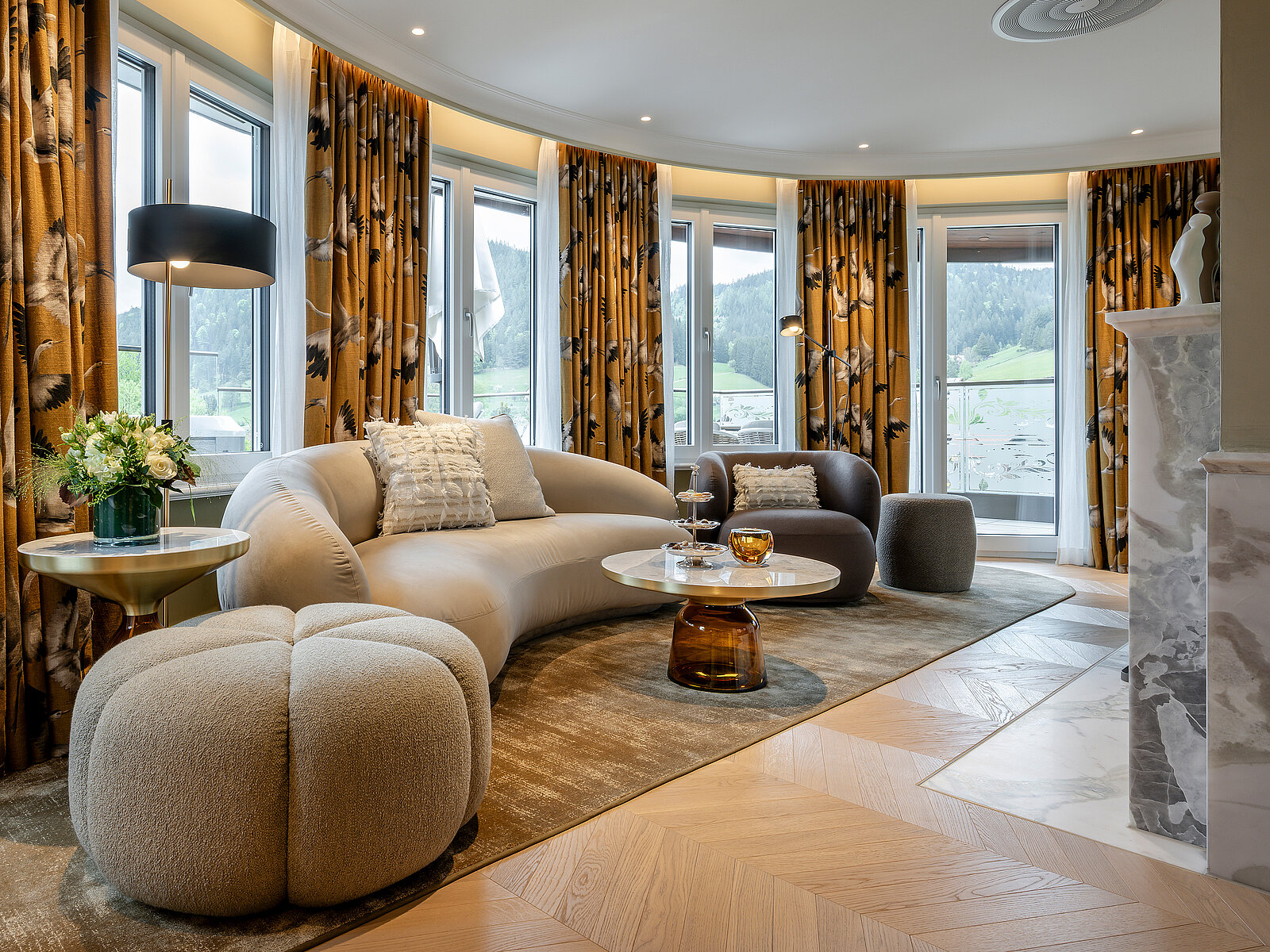 Ein moderner Wohnraum mit großen Fenstern, die einen Blick auf die Landschaft bieten. Eine bequeme, hellbeige Couch steht neben einem dunklen Sessel. Ein runder Tisch ist mit Blumen und einem Getränk dekoriert. Die Gardinen sind floral gemustert und verleihen dem Raum Wärme.