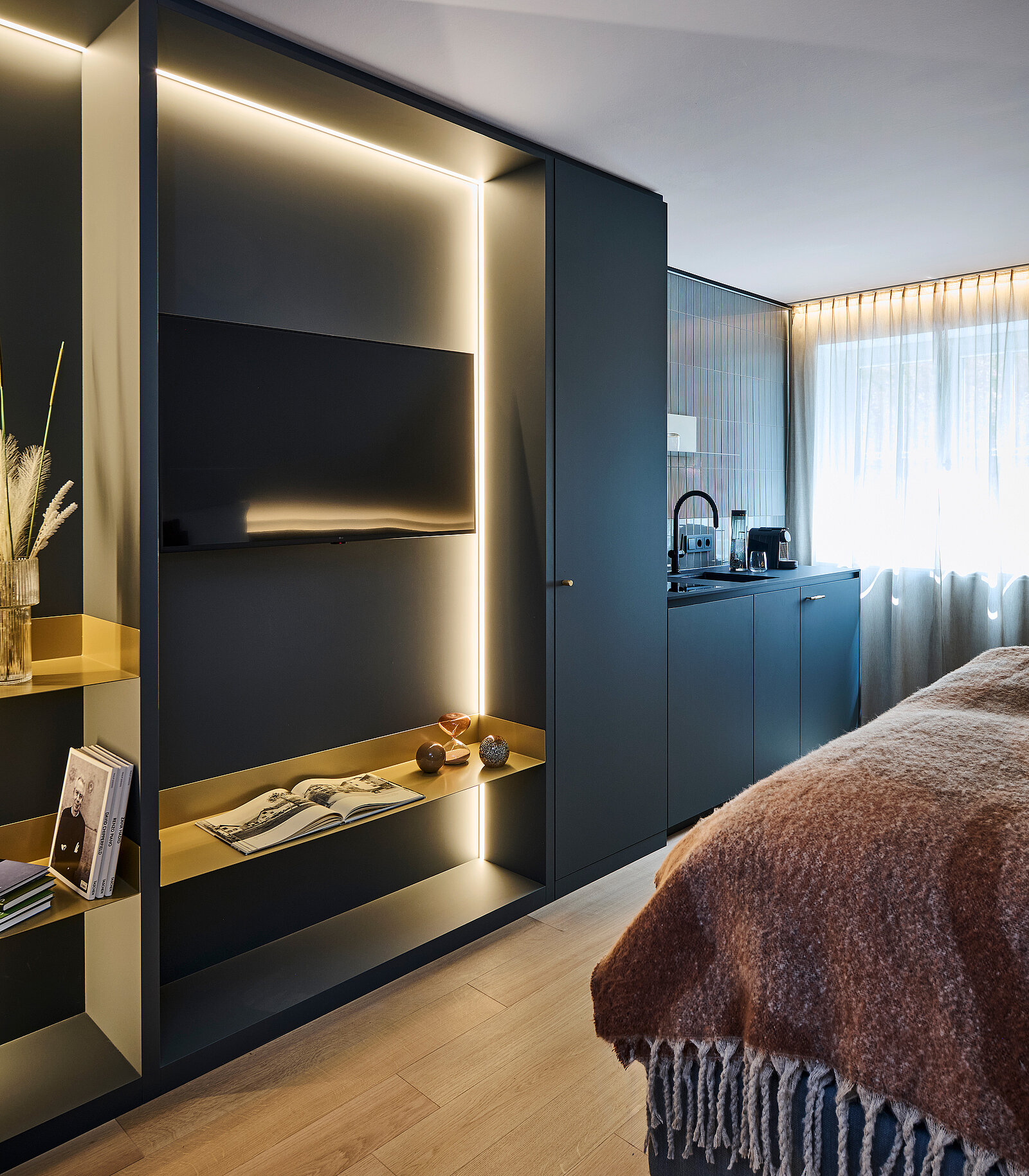 Ein modernes Hotelzimmer mit elegantem Design: Ein eingebauter TV mit Beleuchtung, daneben Regale mit Dekorationen und einem Buch. Eine kleine Küchenzeile mit Spüle und Kaffeemaschine. Ein gemütliches Bett mit einer braunen Decke im Vordergrund.