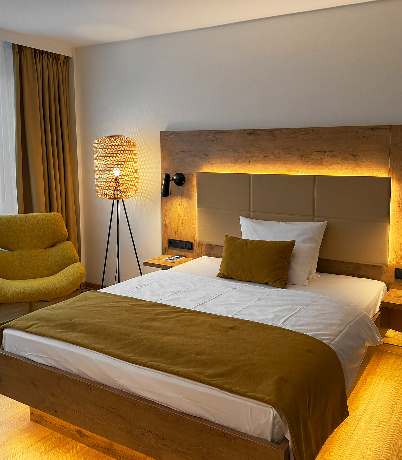 Ein modernes Schlafzimmer mit einem Doppelbett und einem Holzrahmen. Auf dem Bett liegt ein senfgelbes Kopfkissen. Neben dem Bett steht ein bequemer grüner Sessel. Eine stylische Lampe mit einem geflochtenen Schirm erleuchtet den Raum. Die Wände sind hell, und es gibt Vorhänge an einem Fenster.