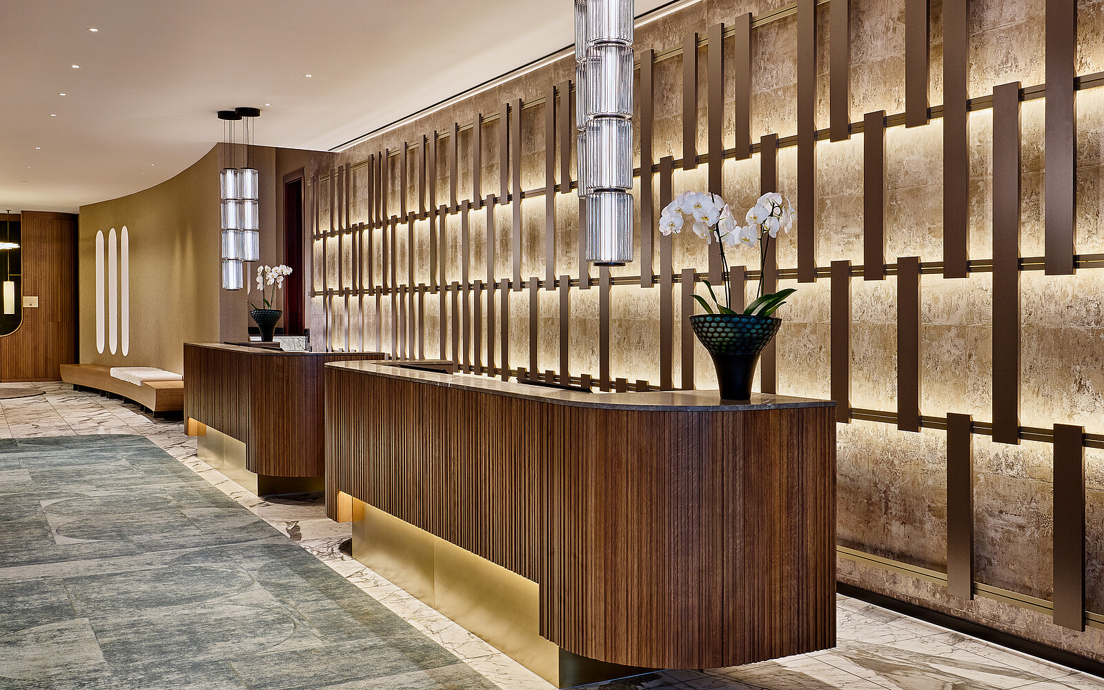 Ein eleganter und moderner Empfangsbereich in einem Hotel, gekennzeichnet durch eine lange, geschwungene Rezeption aus Holz. Rechts befindet sich eine beleuchtete Wand mit stilvollen Musterelementen, während links auf der Theke eine Vase mit weißen Orchideenblüten steht.