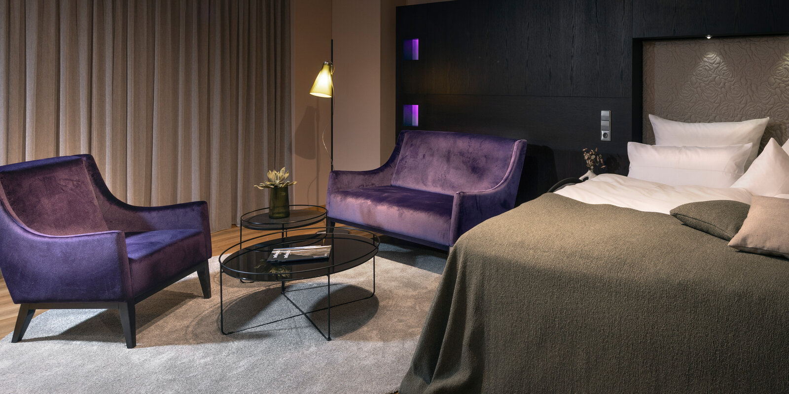Modern hotel room with a large bed, purple armchairs, round glass tables, and soft lighting. Draped curtains and a small plant add elegance.