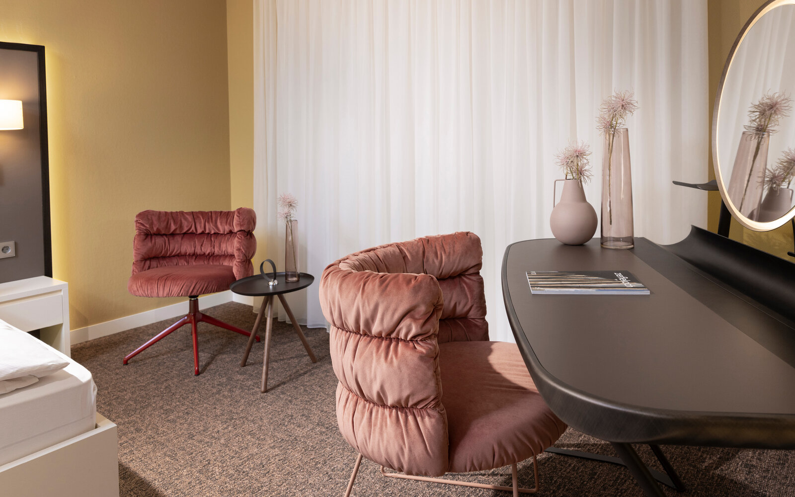 Modern hotel room with pink cushioned chairs, a sleek black desk, round mirror, and soft lighting. Neutral tones and minimalist decor.