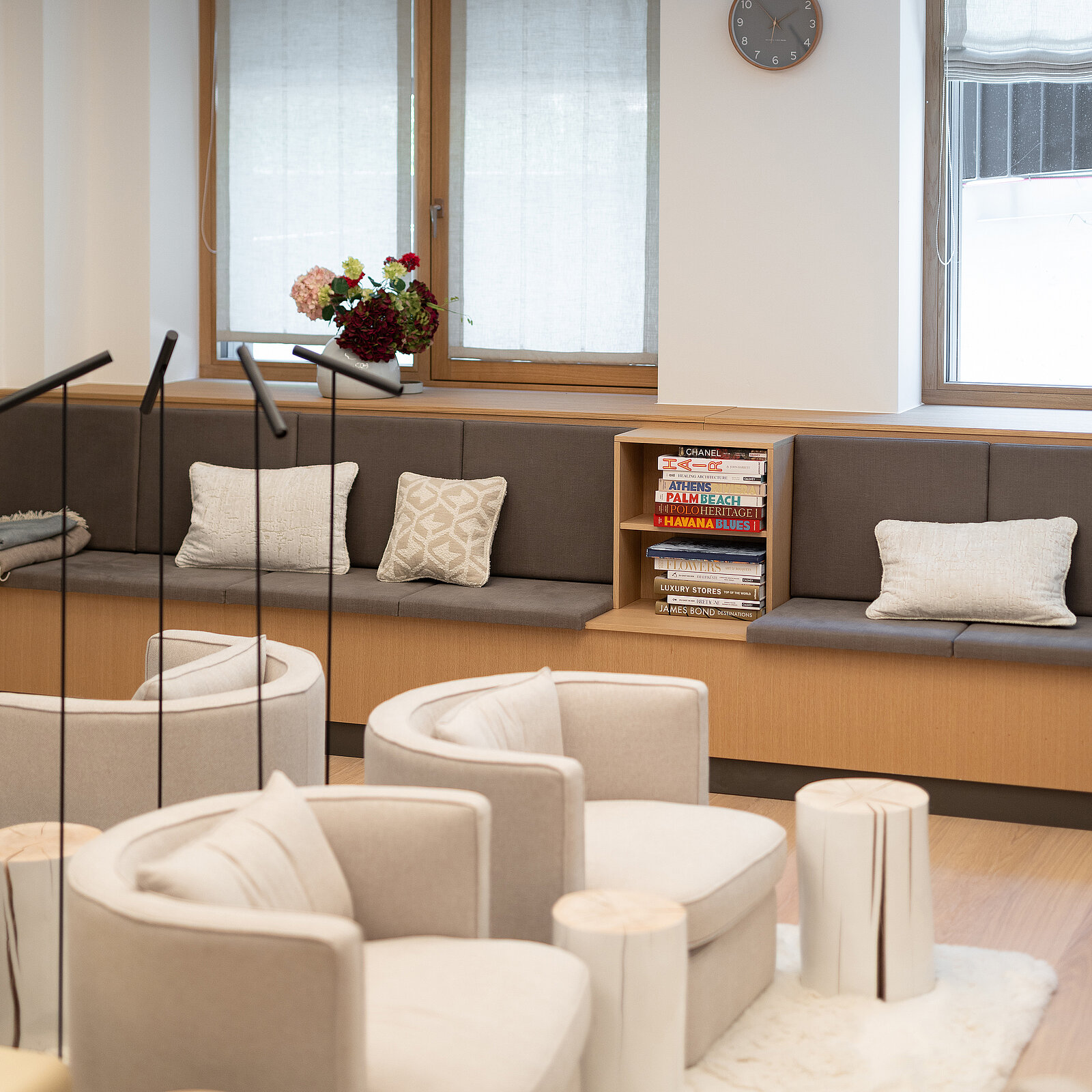 Gemütlicher Warteraum mit hellen Sesseln, modernem Design und Kissen auf einer langen Sitzbank. Ein Regal mit Büchern und Dekoration steht neben den Fenstern. Eine Uhr hängt über den Fenstern, die mit halbtransparenten Vorhängen bedeckt sind.